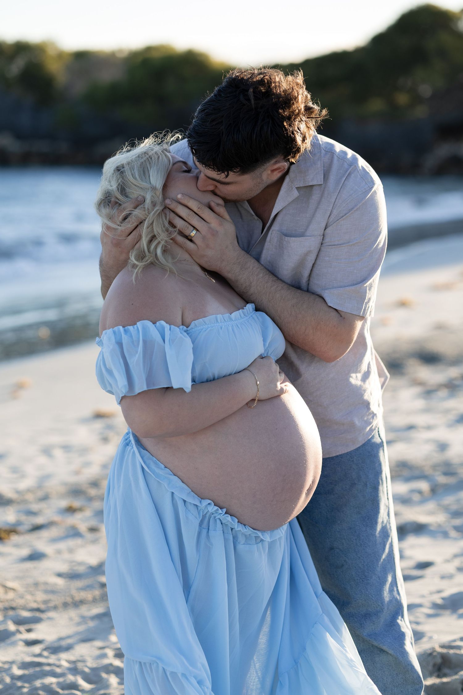 Pregnant woman in blue off-shoulder dress kissing partner on sandy beach at sunset. Woodman Point Perth