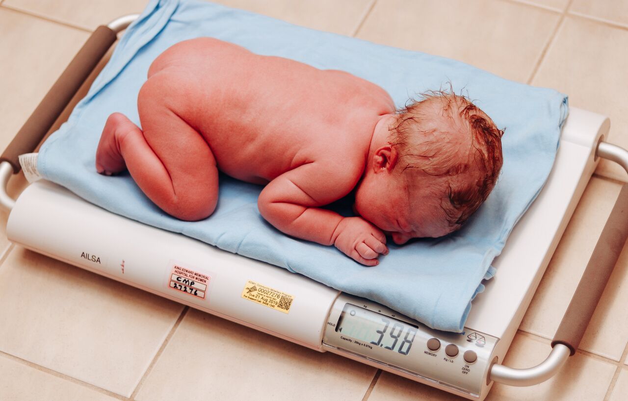 Newborn baby lying on a blue blanket on a digital scale showing weight measurement in a hospital.