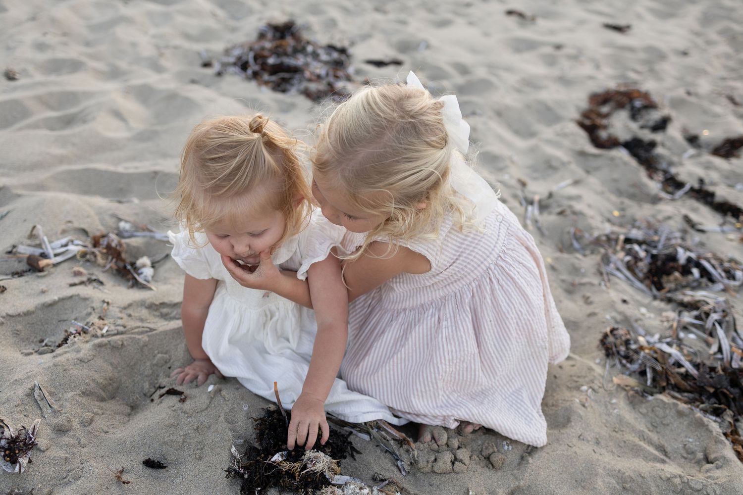 Two young blonde girls in white dresses playing together on a sandy beach near seaweed. Cape Peron Perth