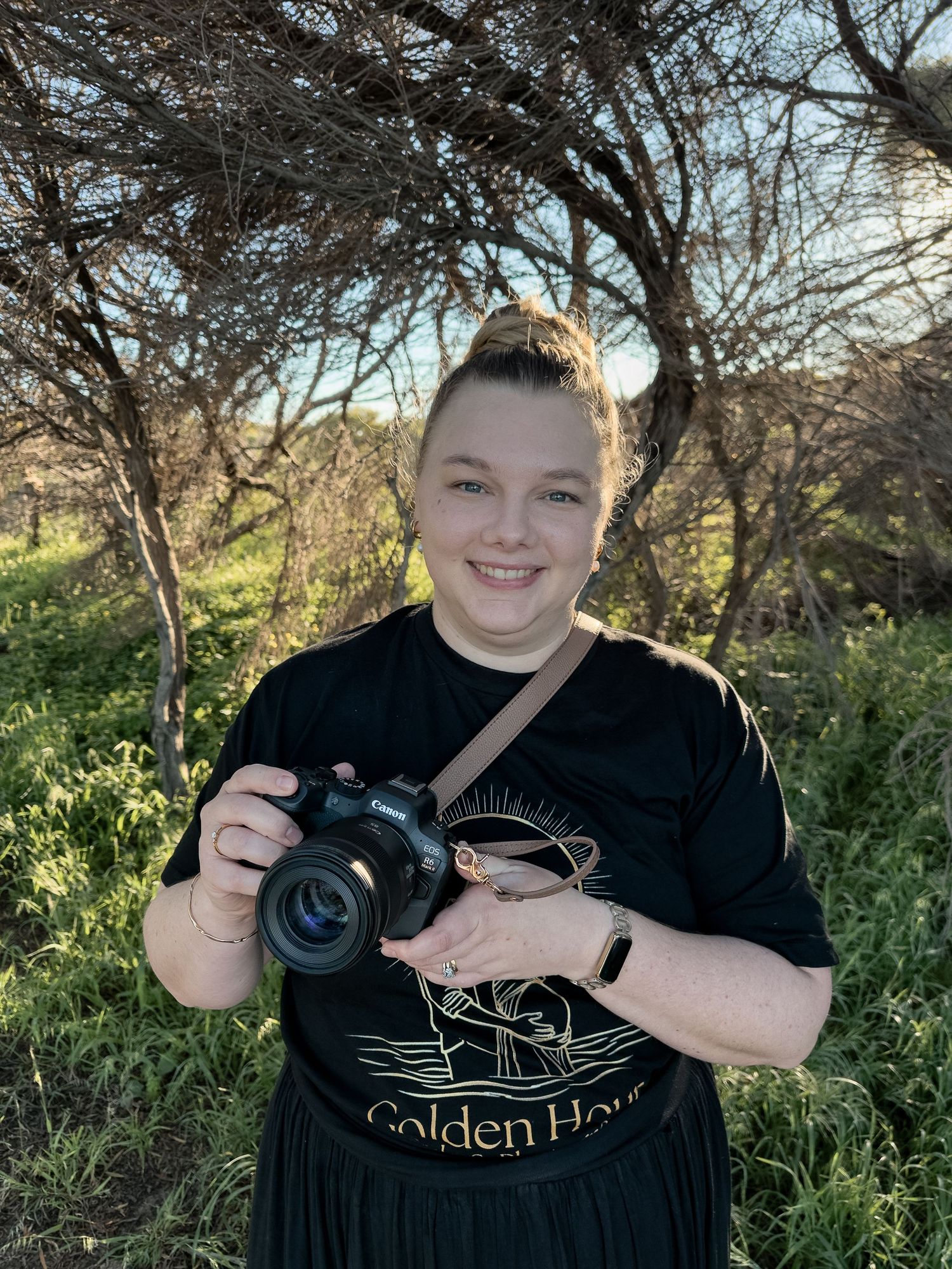 Woman smiling outdoors holding a camera, wearing a black Golden Hour t-shirt, surrounded by trees and greenery.
