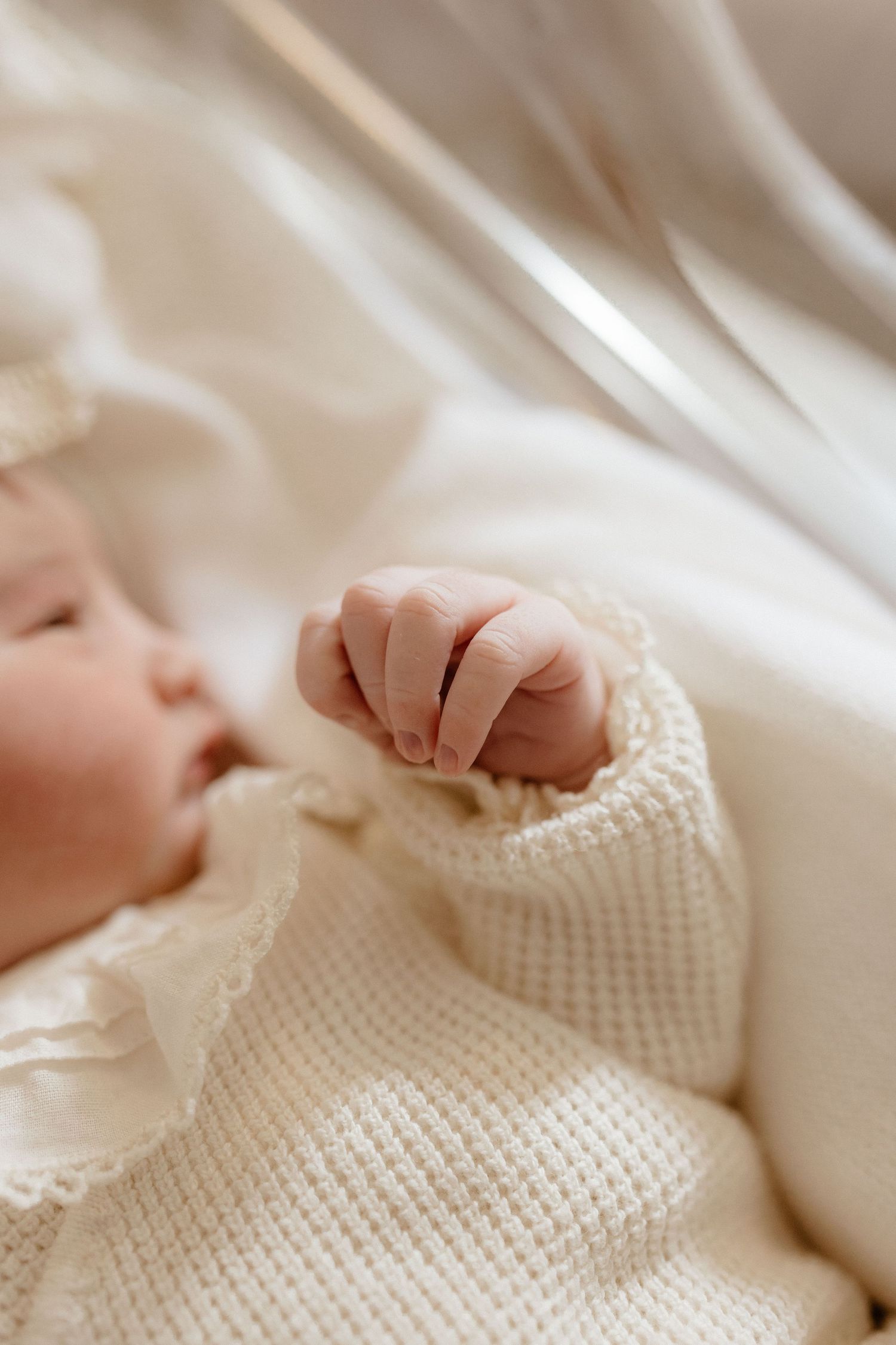 Close-up of sleeping newborn baby's tiny clenched fist in cream knit outfit on soft bedding.