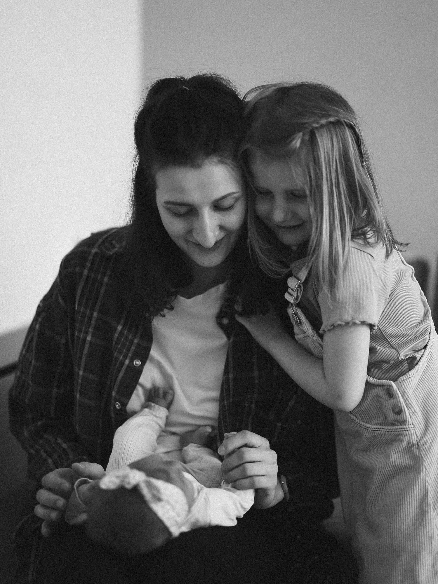 Black and white portrait of mother holding newborn baby with young daughter leaning close.