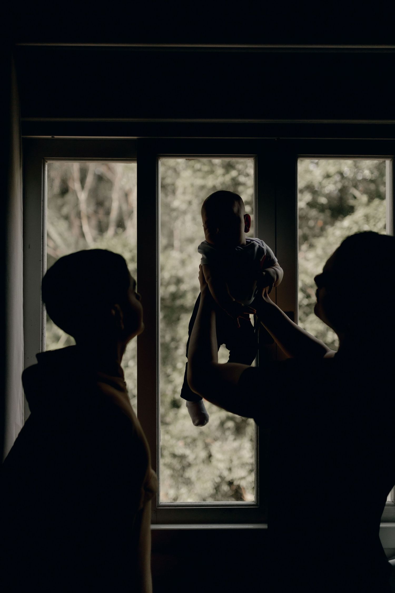 Silhouette of two parents lifting baby near a window with natural light.