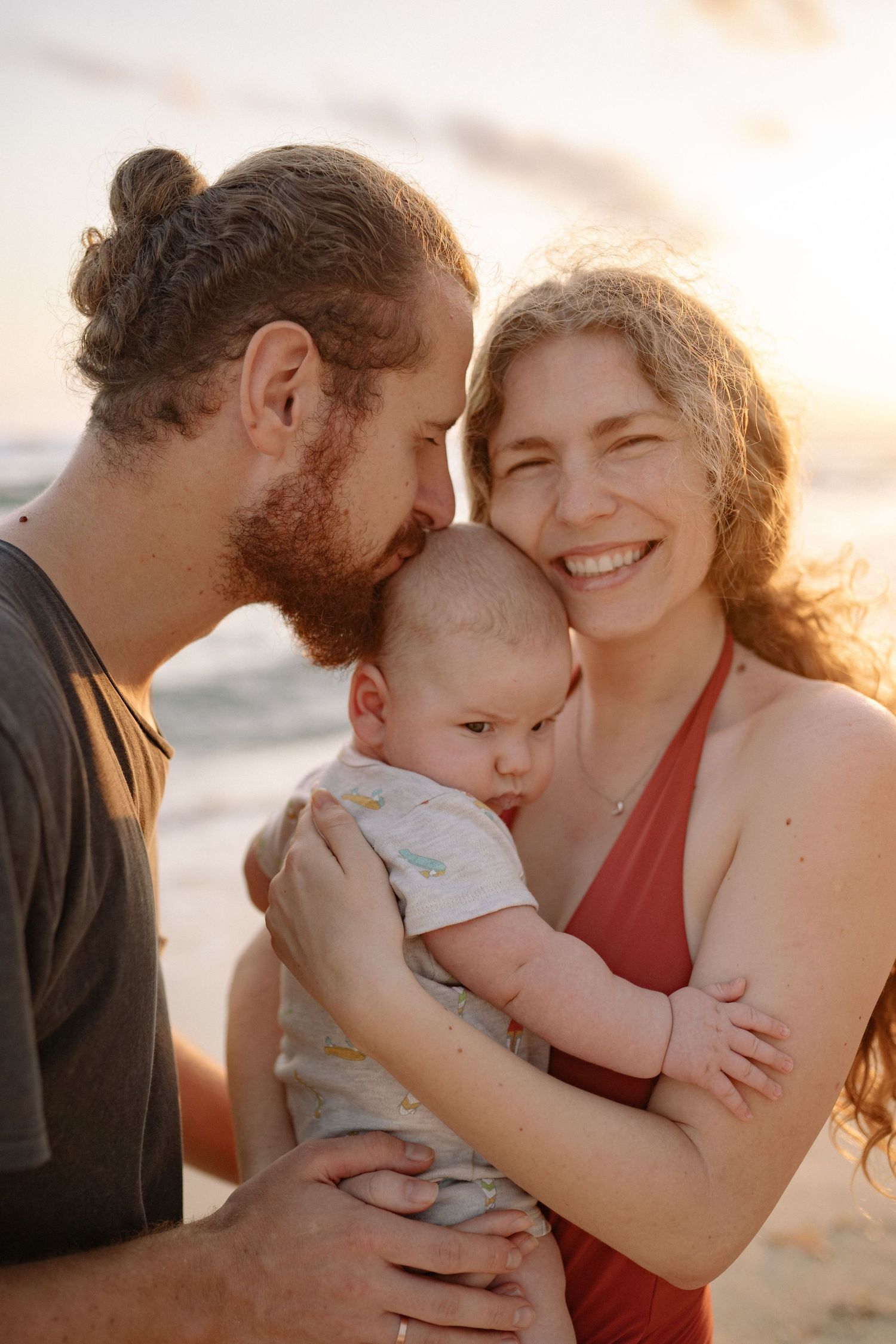 Couple with newborn baby sharing a tender moment on the beach at golden hour.