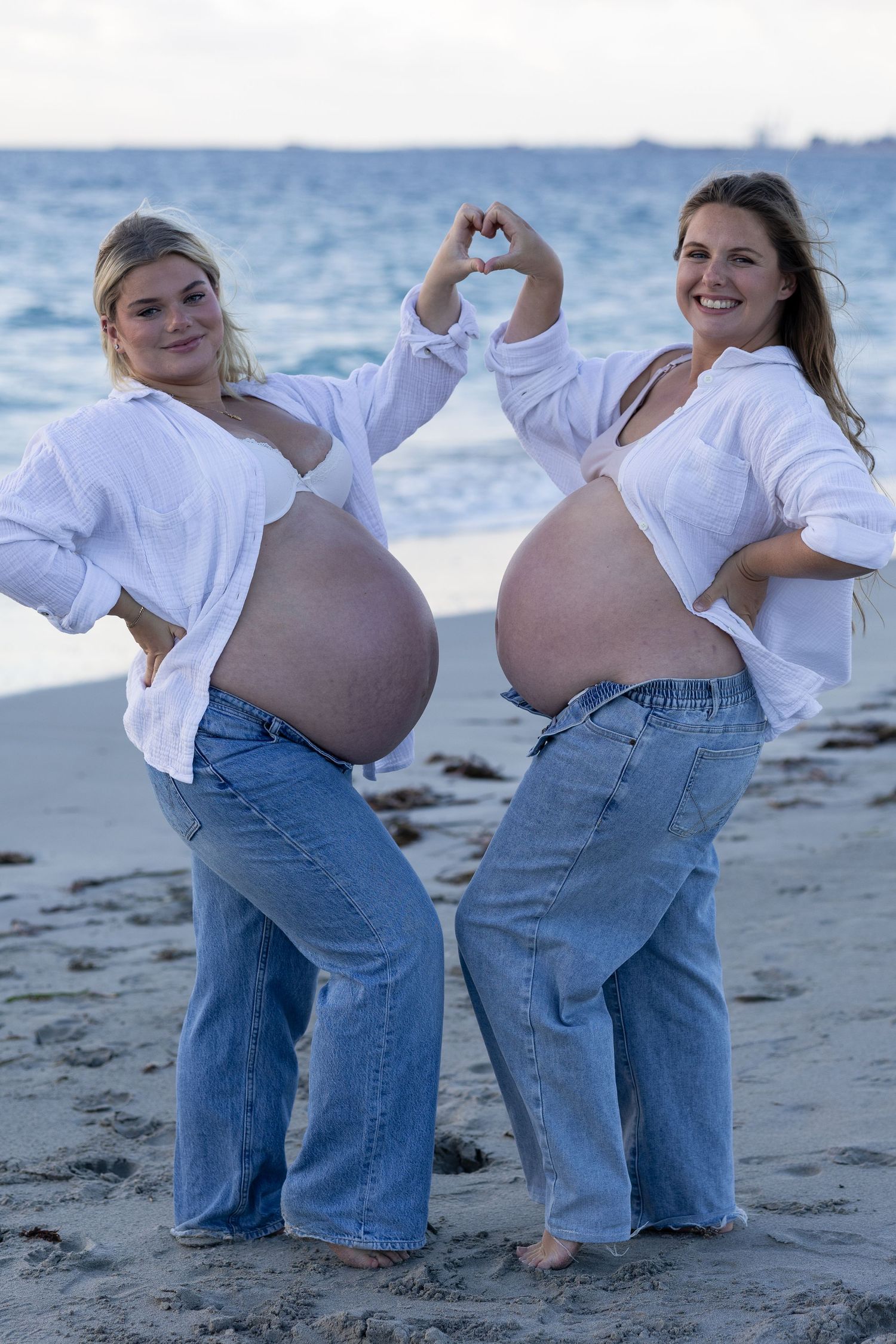 Two pregnant women in white shirts and jeans show their baby bumps on a beach, one forming a heart shape with her hands.