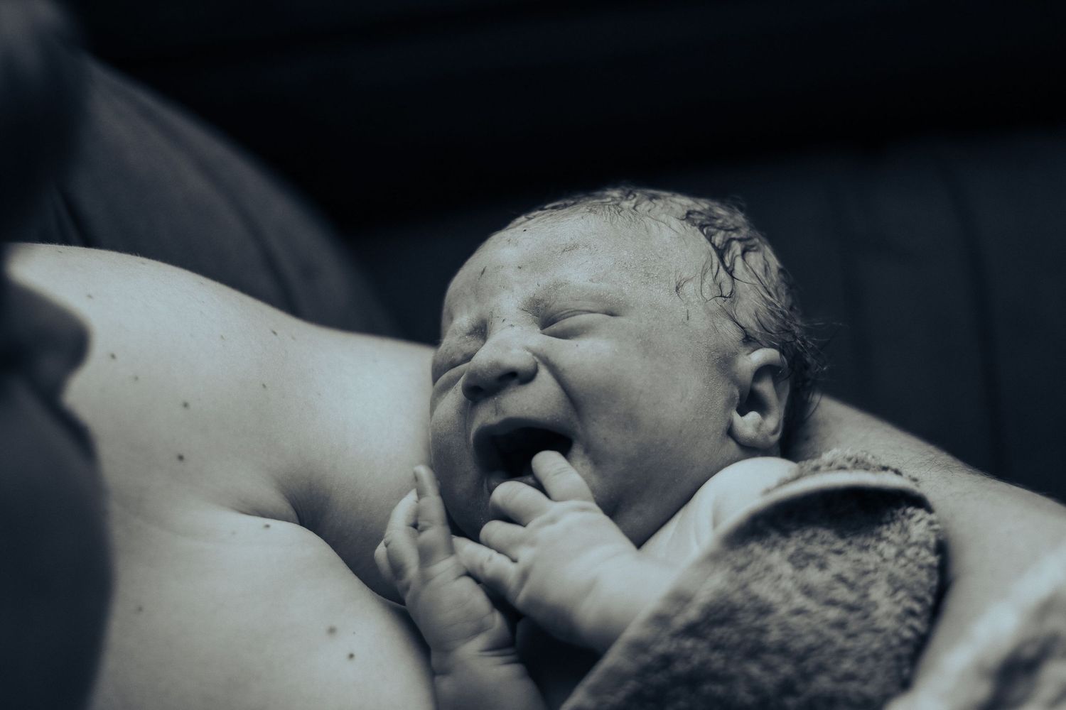 Black and white photo of a newborn baby yawning moments after birth, held skin-to-skin by mother.