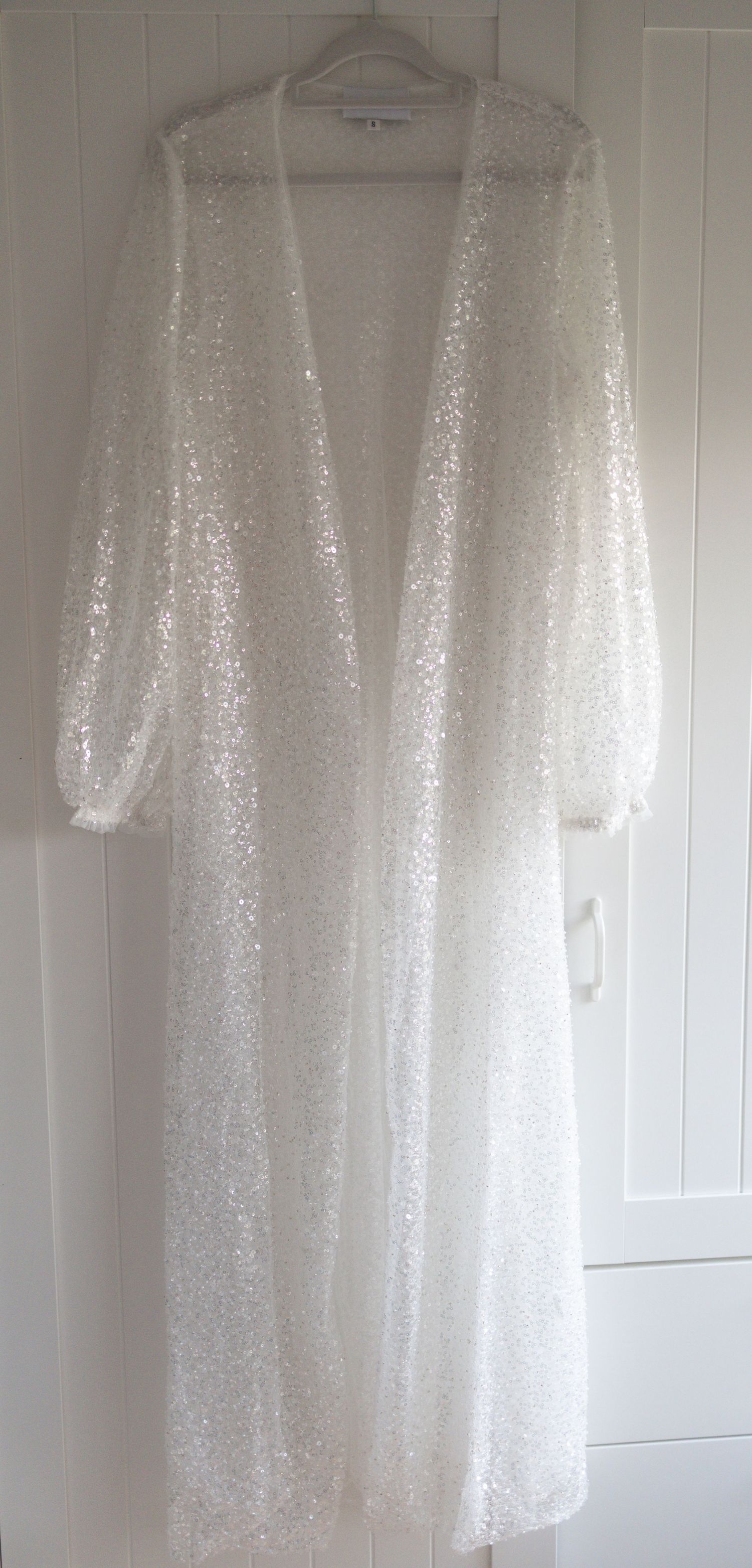 White sequin maxi duster coat with deep V-neckline, balloon sleeves, and full-length open front design, hanging on display.