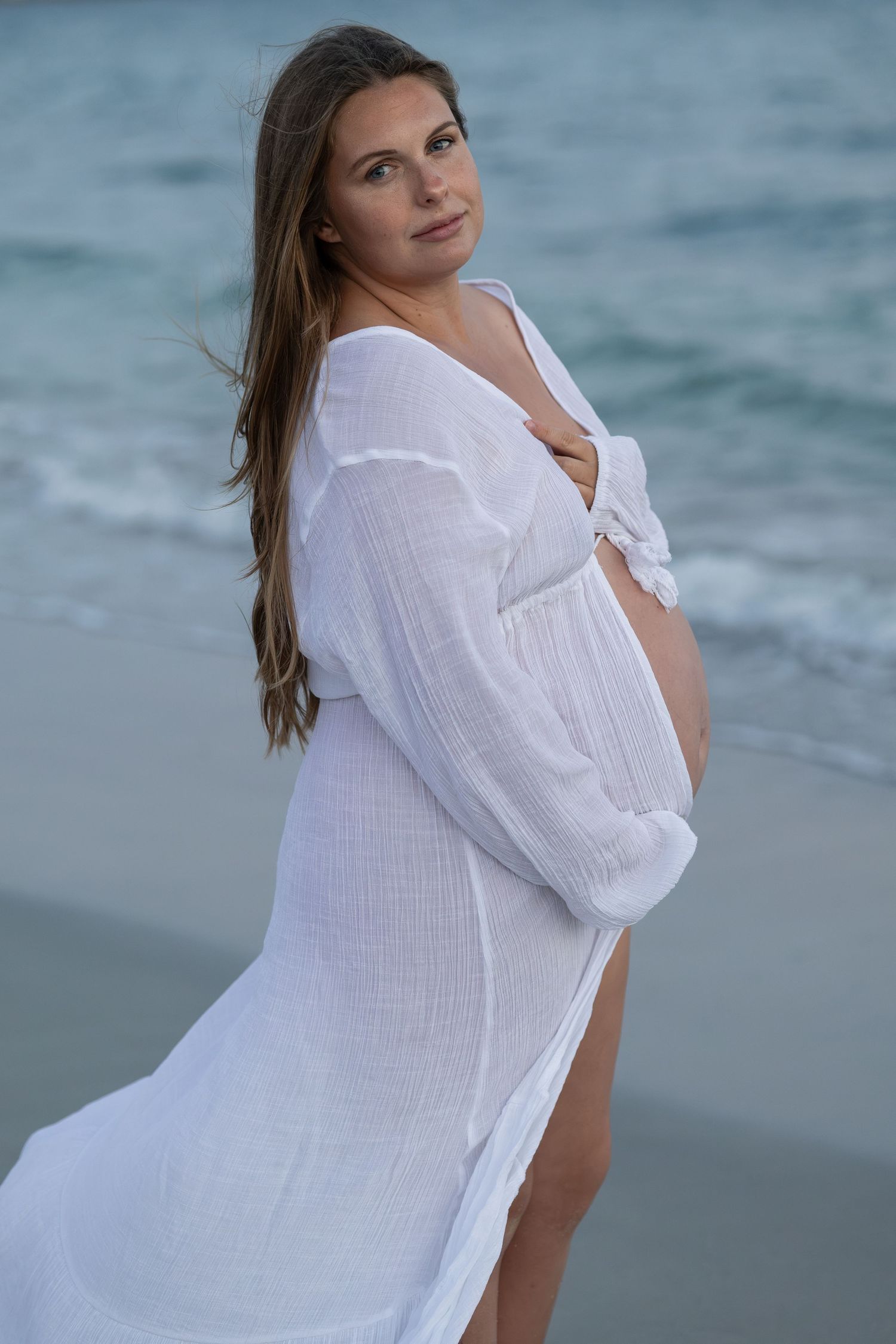Pregnant woman in flowing white dress posing on beach, holding baby bump with ocean waves behind her. Woodman Point Perth