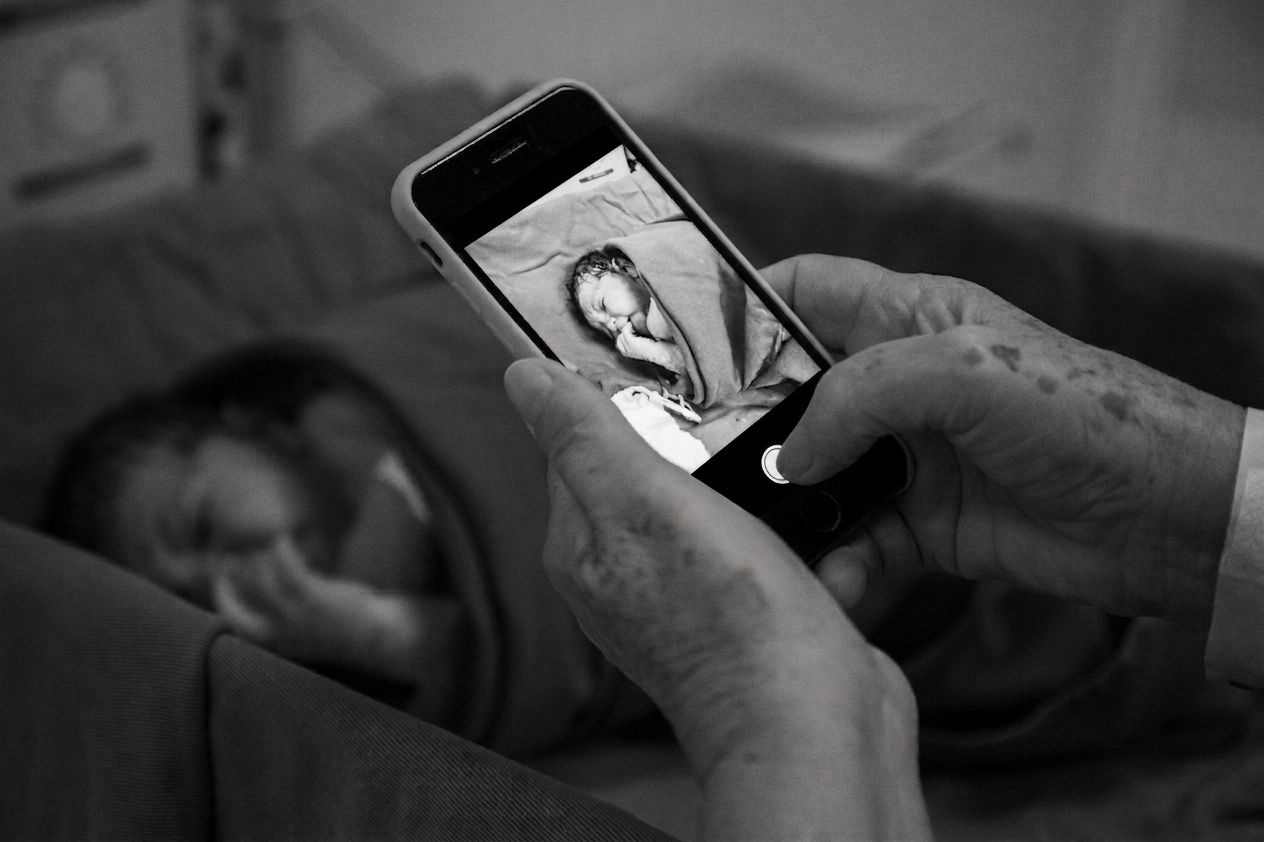 Black and white photo of hands photographing a newborn baby in a hospital bassinet with a smartphone.