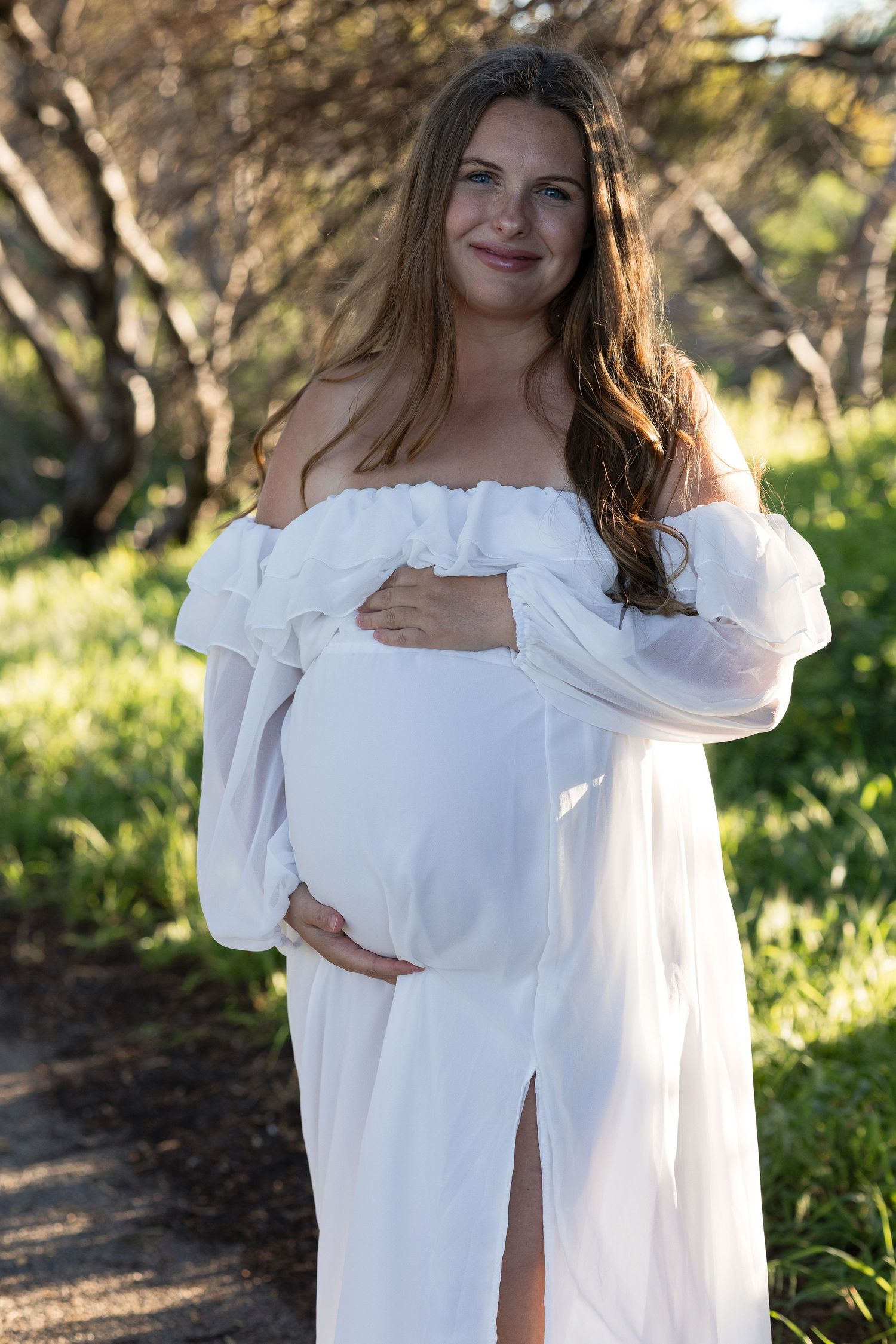 Smiling pregnant woman in white off-shoulder dress cradling baby bump outdoors among green trees.