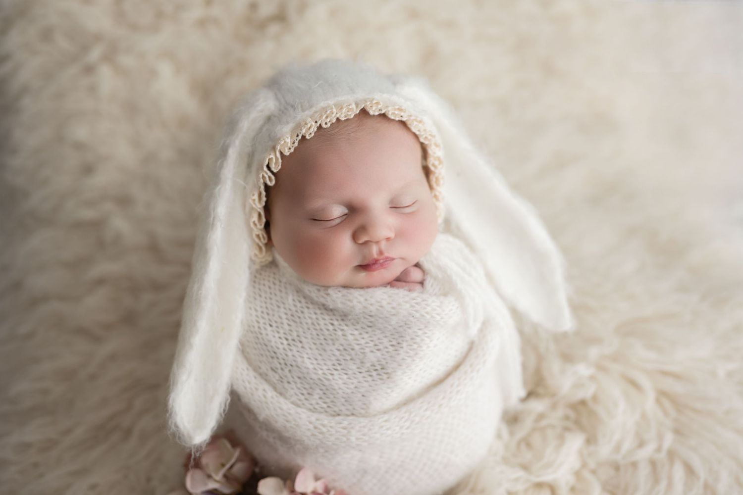 Sleeping newborn baby wrapped in white knit swaddle wearing bunny ear bonnet on fluffy rug.