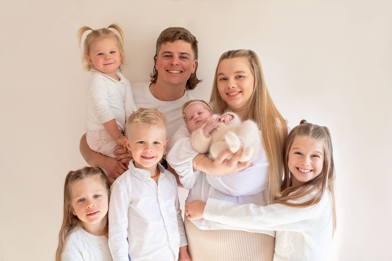 Happy family of seven dressed in white posing together with their newborn baby at home.