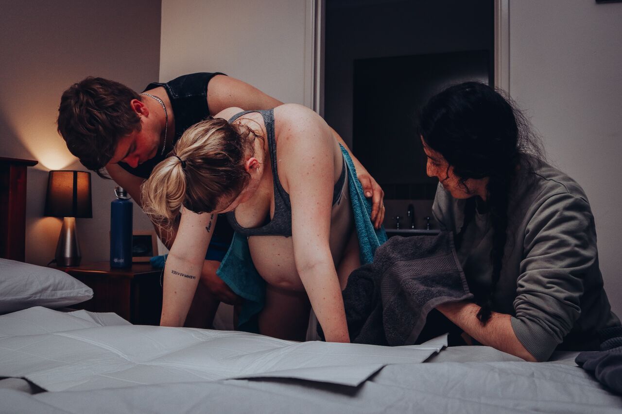 Birth support team assisting a laboring woman leaning over a bed during active home labor.