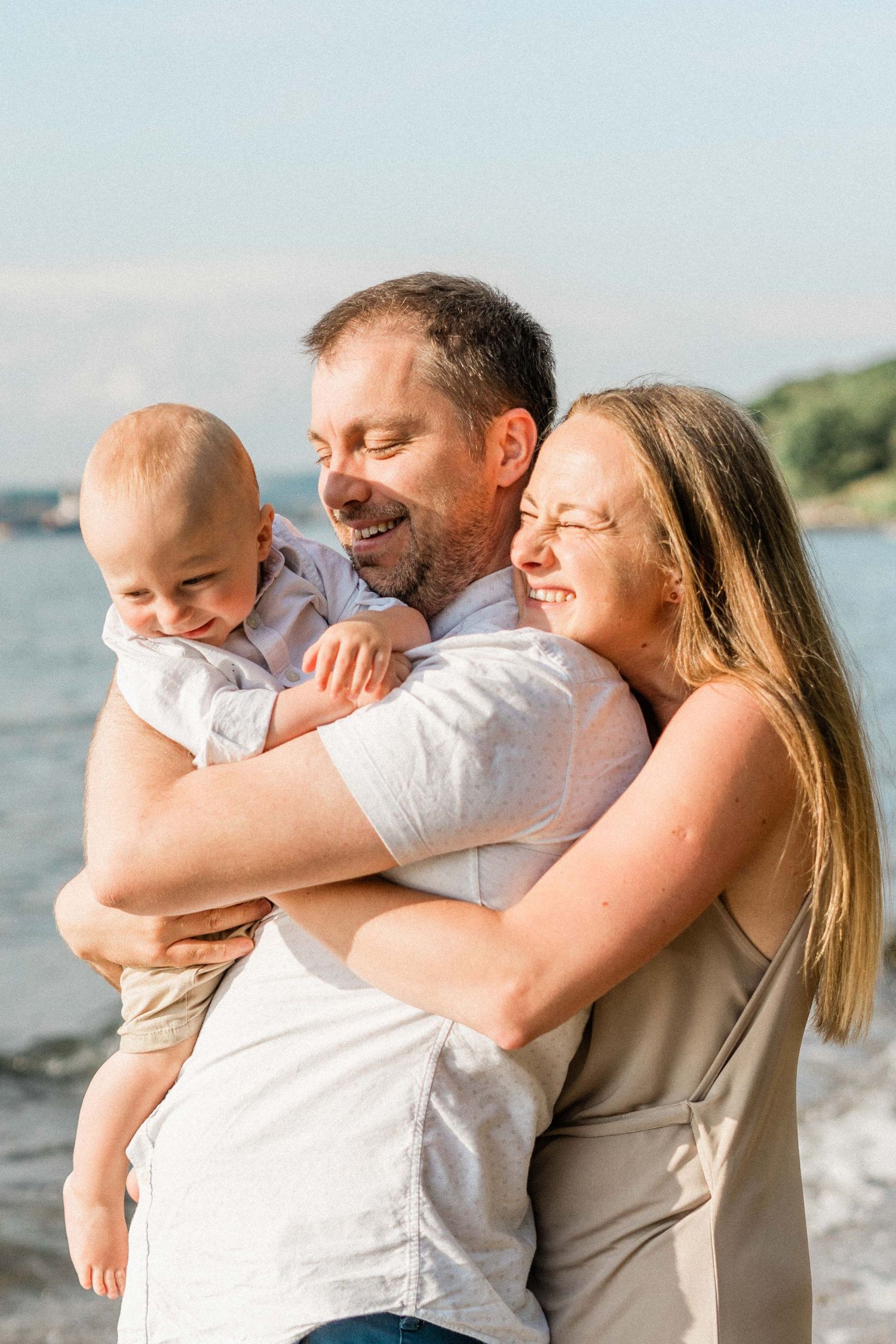 Happy family with baby smiling and hugging near the ocean on a sunny day.