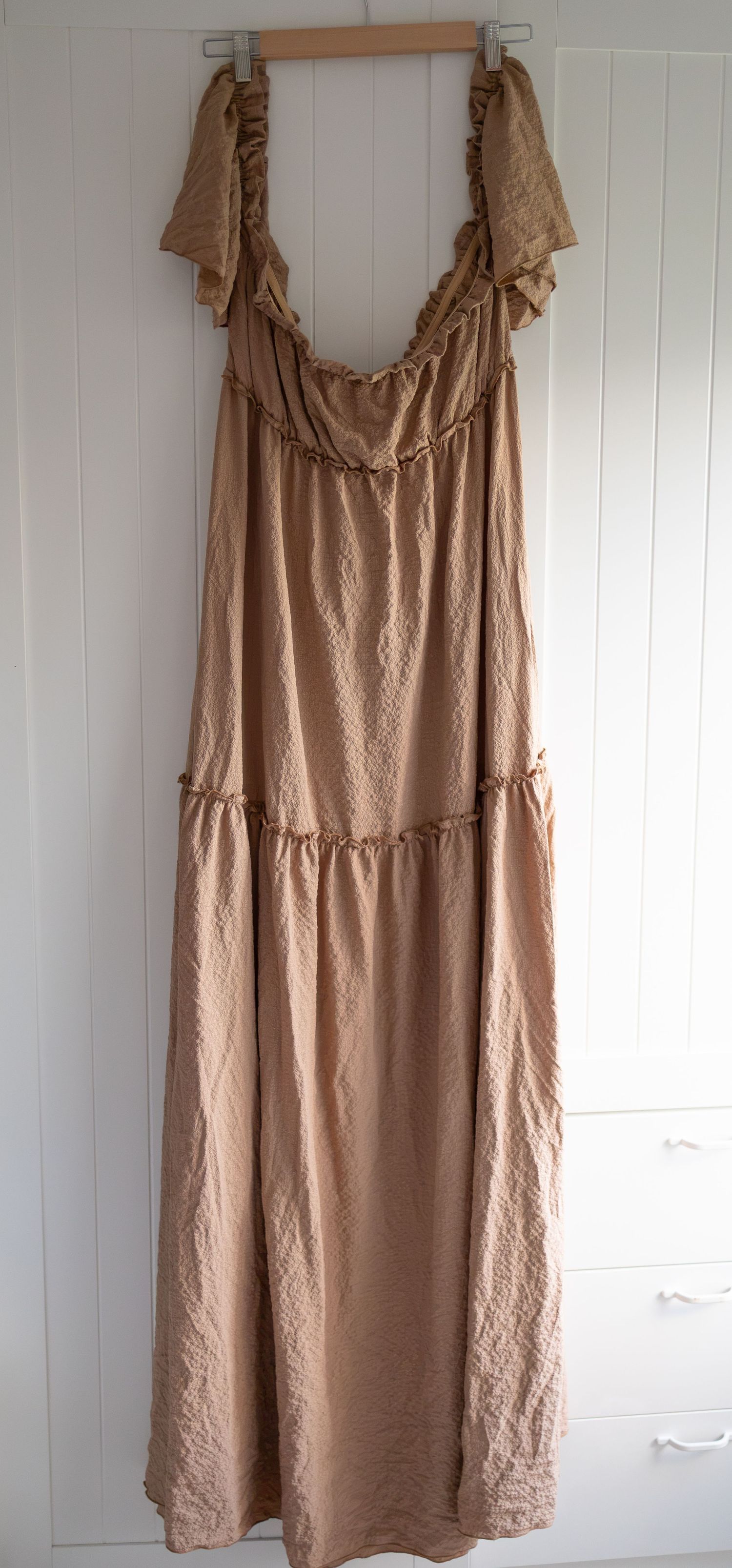 Beige ruffle maxi dress on hanger, sleeveless tiered design with delicate frills, soft neutral tone, floor-length silhouette.