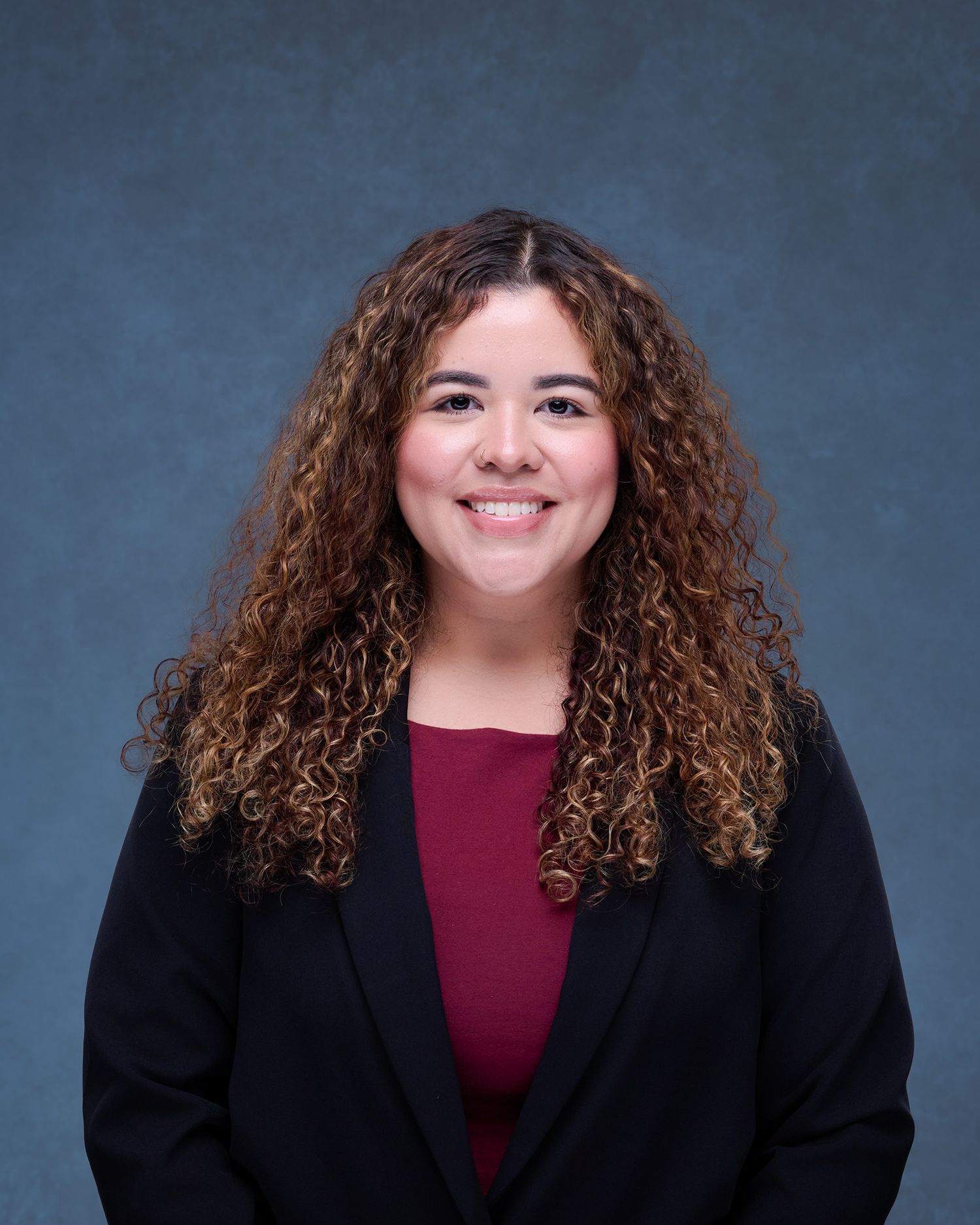 Professional headshot of a smiling woman with curly auburn hair wearing a black blazer and burgundy top.