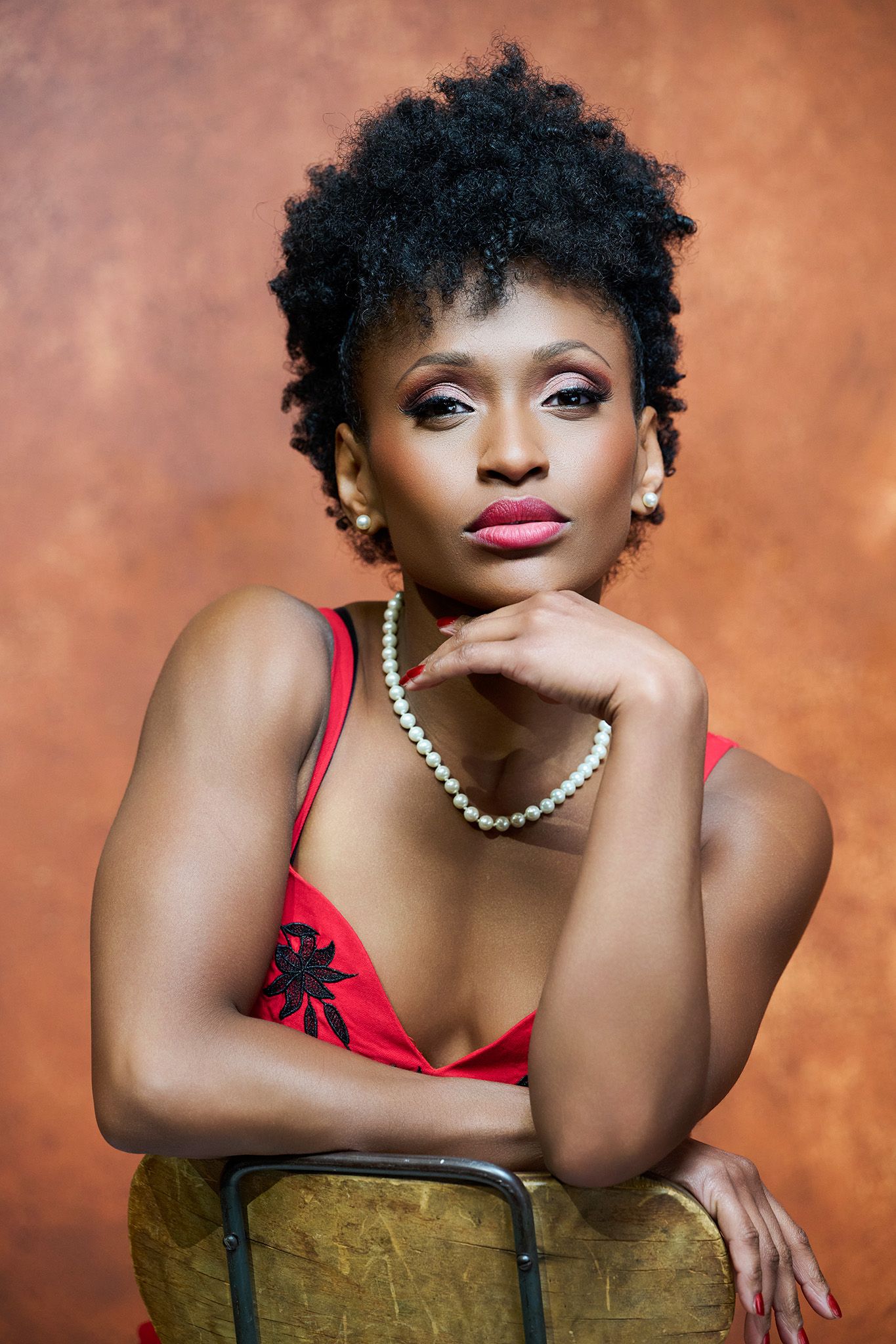 An elegant woman in a red dress and pearl necklace poses with natural updo hair on chair.