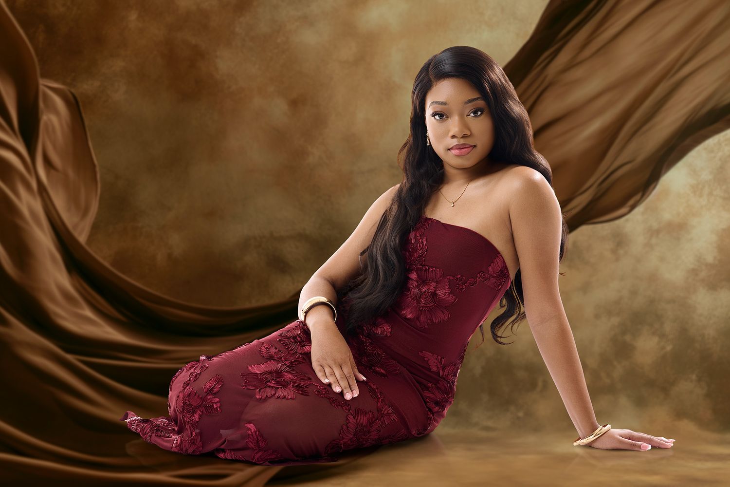 Young woman in elegant burgundy lace gown poses gracefully against a flowing brown fabric studio backdrop.