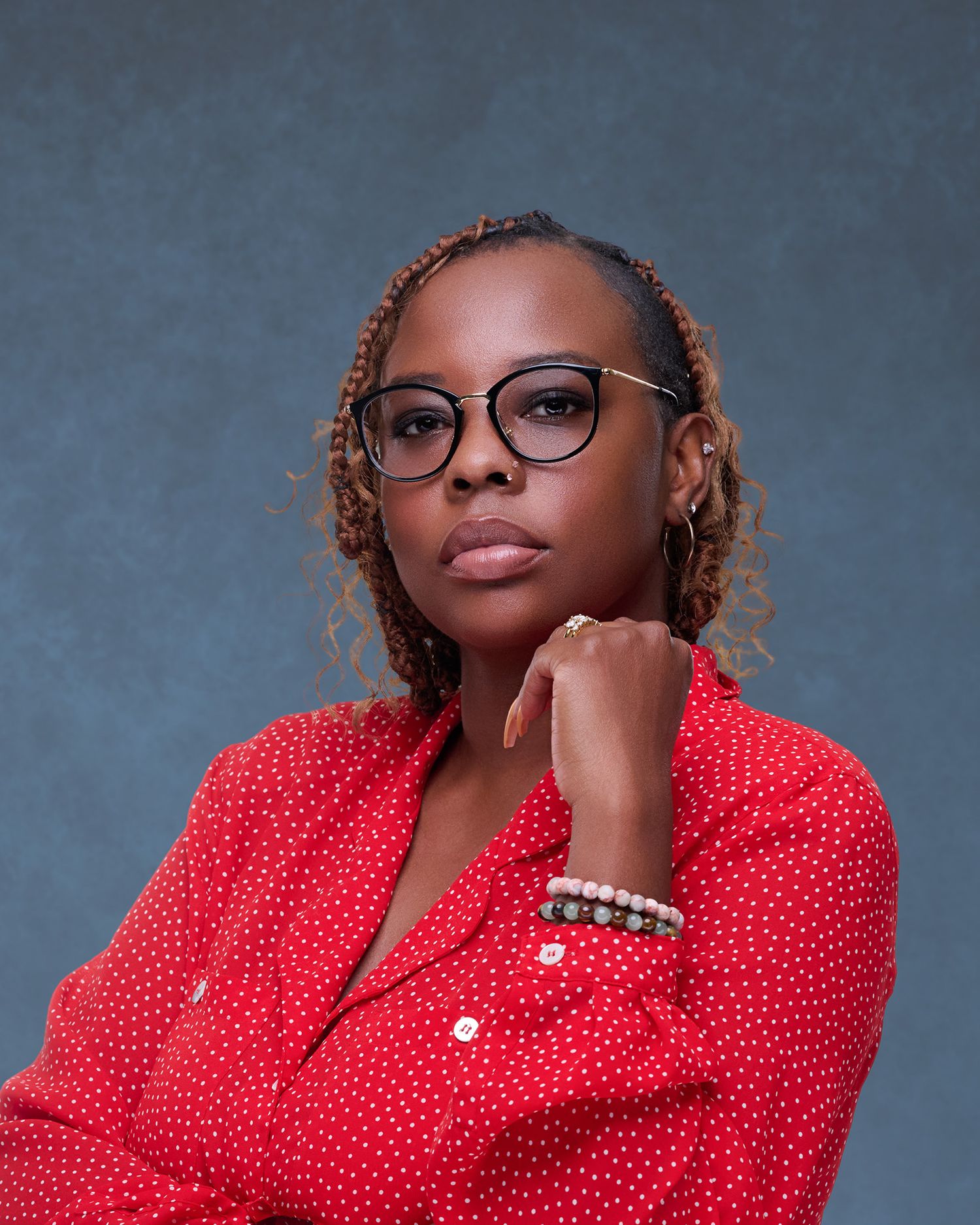 Woman in red polka dot shirt and glasses poses confidently against gray background with braided hair and pearl bracelet.