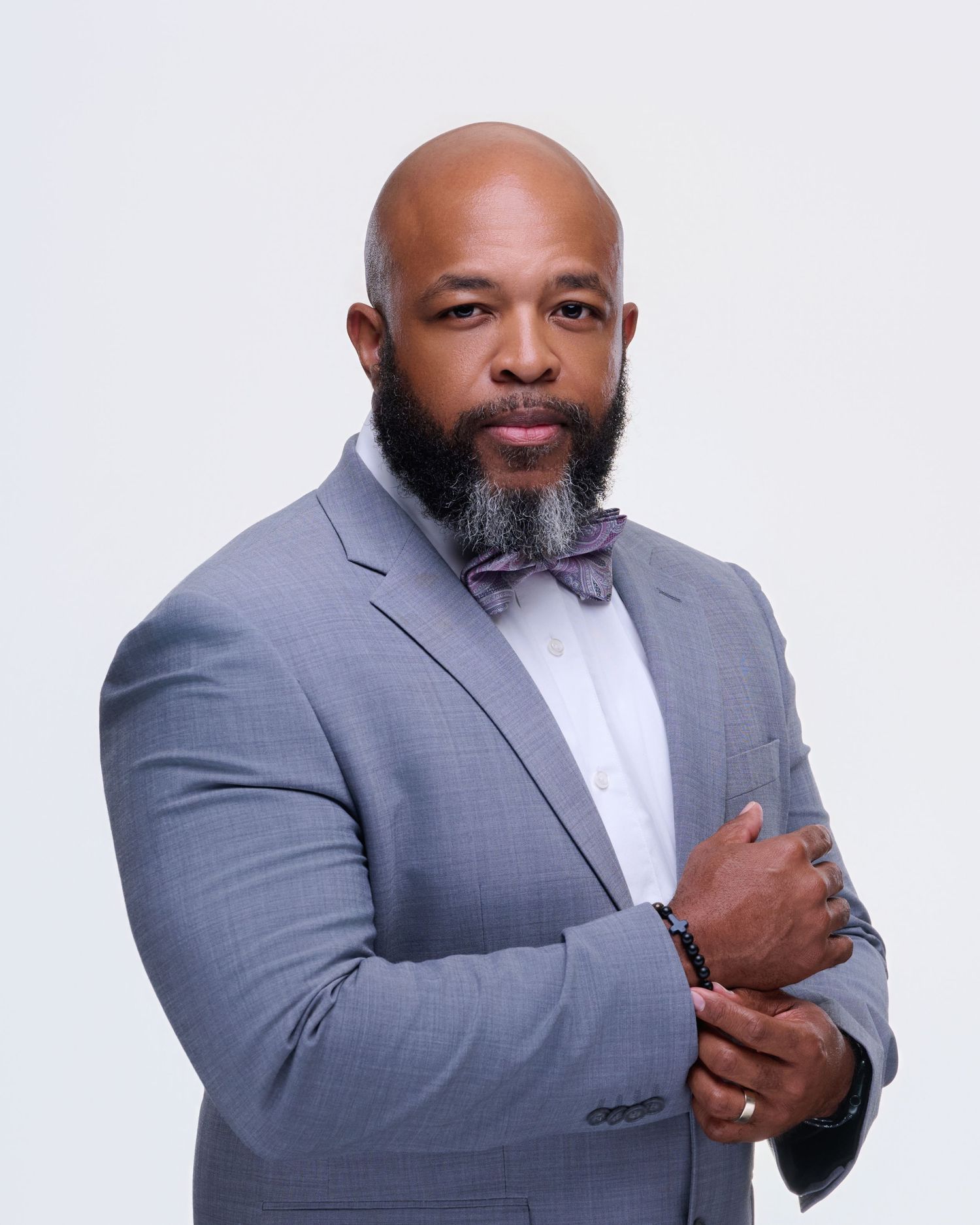 Bald Black man with gray beard wearing gray suit and purple bow tie poses confidently against white background.