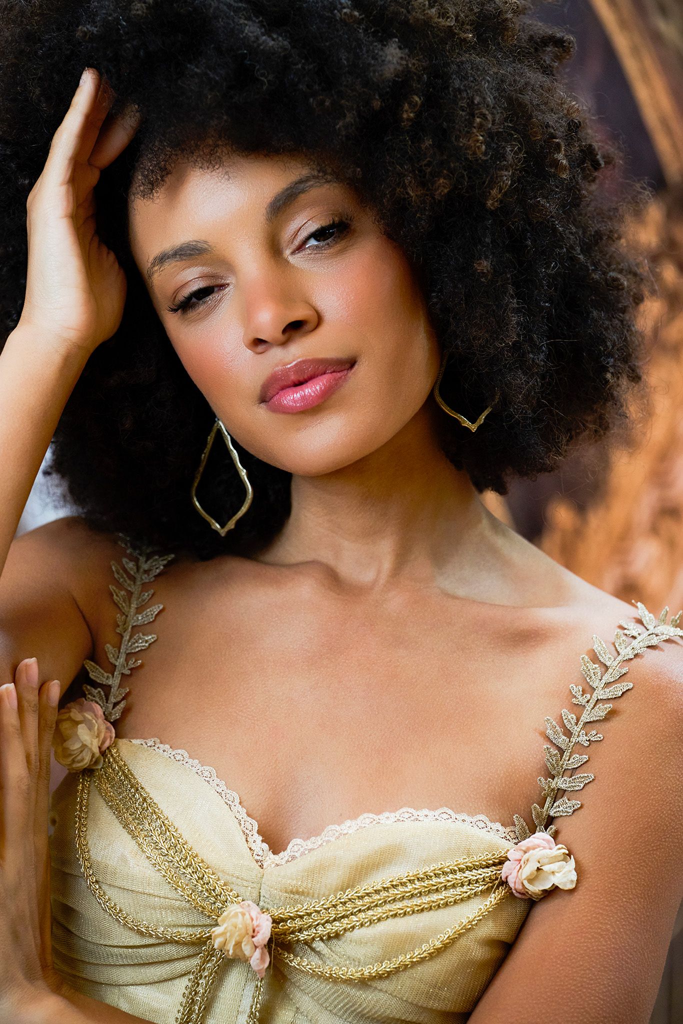 A woman with a natural afro hairstyle poses in a gold lace corset top with leaf-shaped gold earrings.