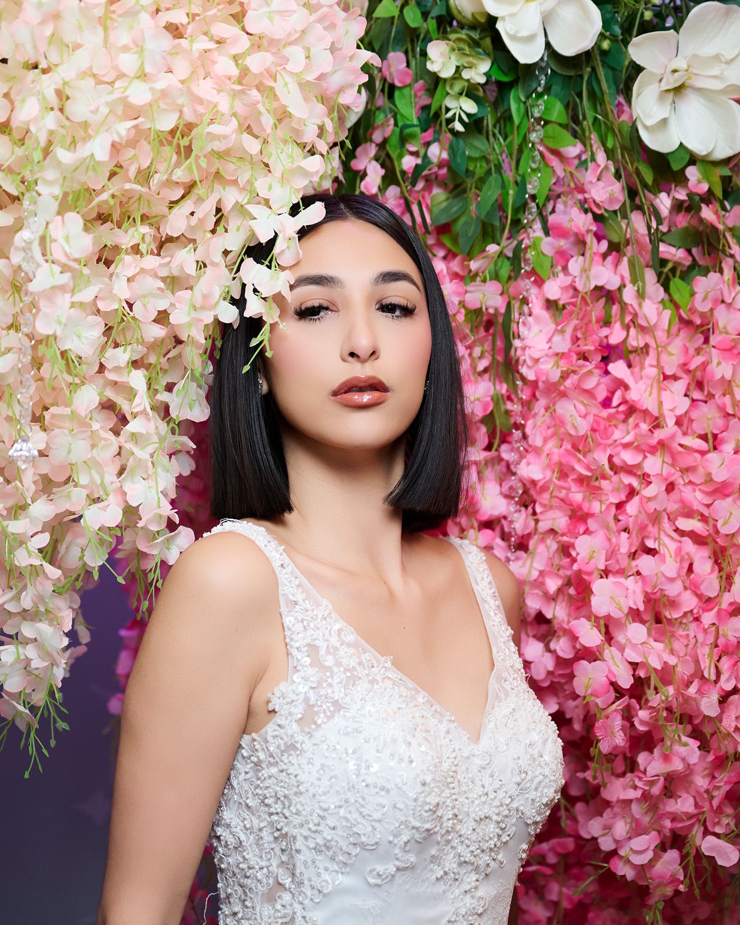 A woman with a black bob poses in a lace bridal gown amid pink and cream flowers.