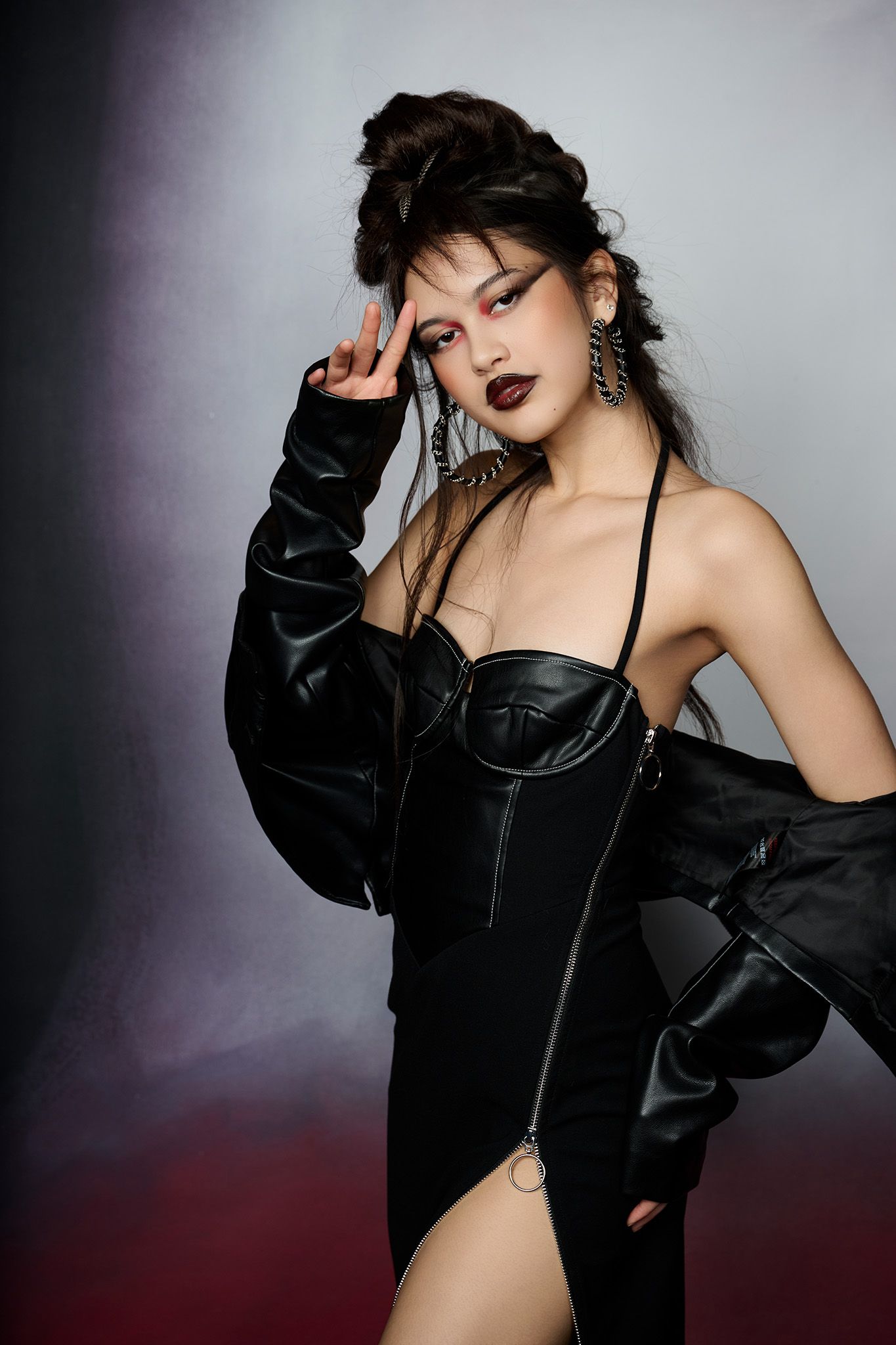 A woman in a black leather corset bodysuit and jacket poses with bold dark makeup in a studio.