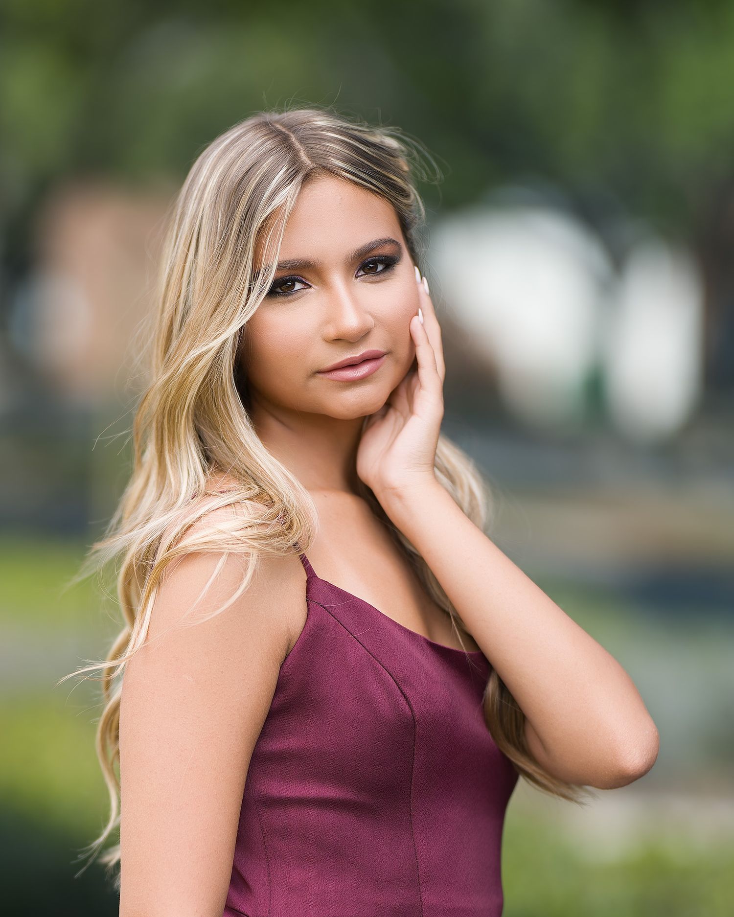 Young blonde woman in burgundy dress posing outdoors with soft bokeh green background.