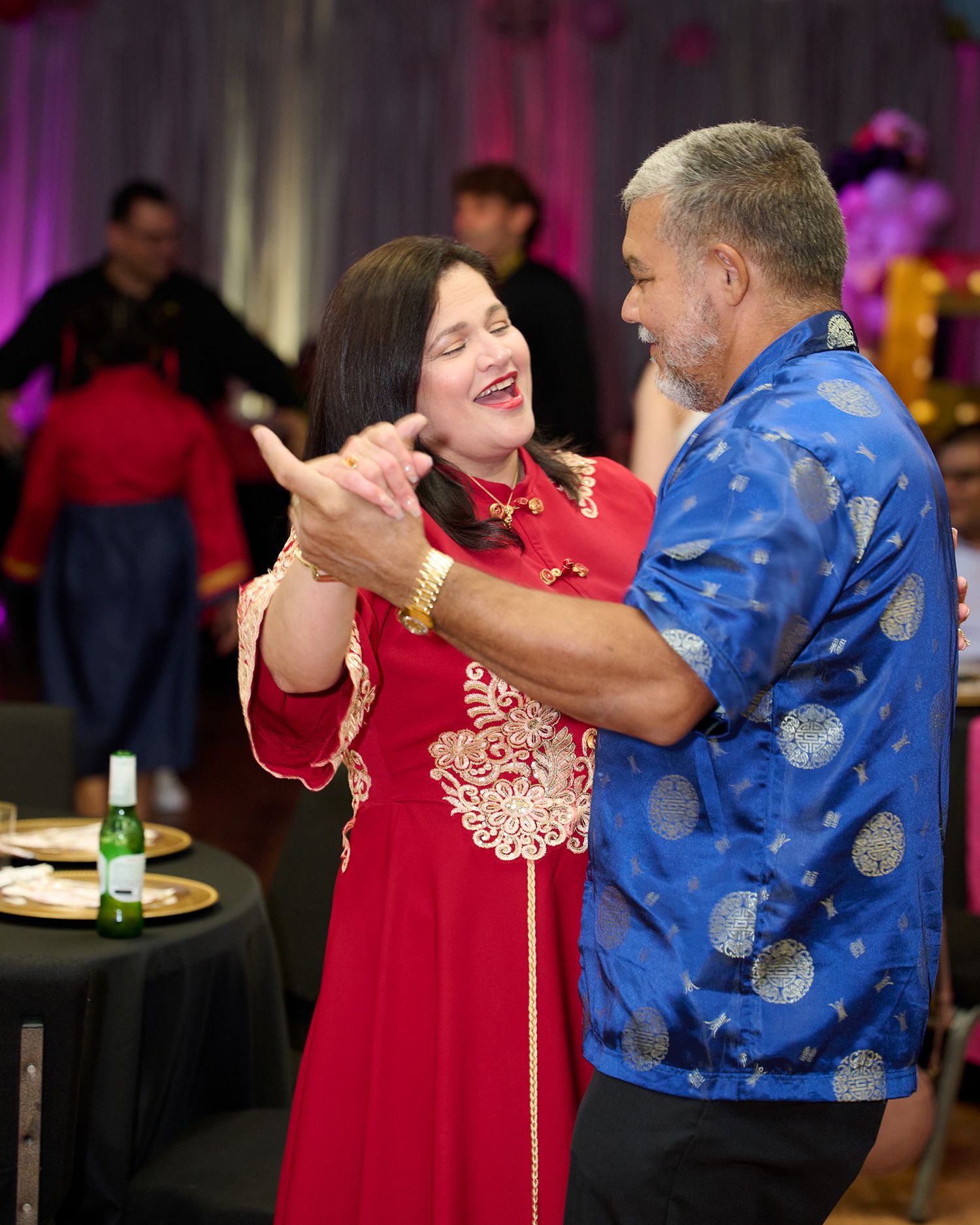 Couple dancing together at a festive event, woman in red dress and man in blue patterned shirt.