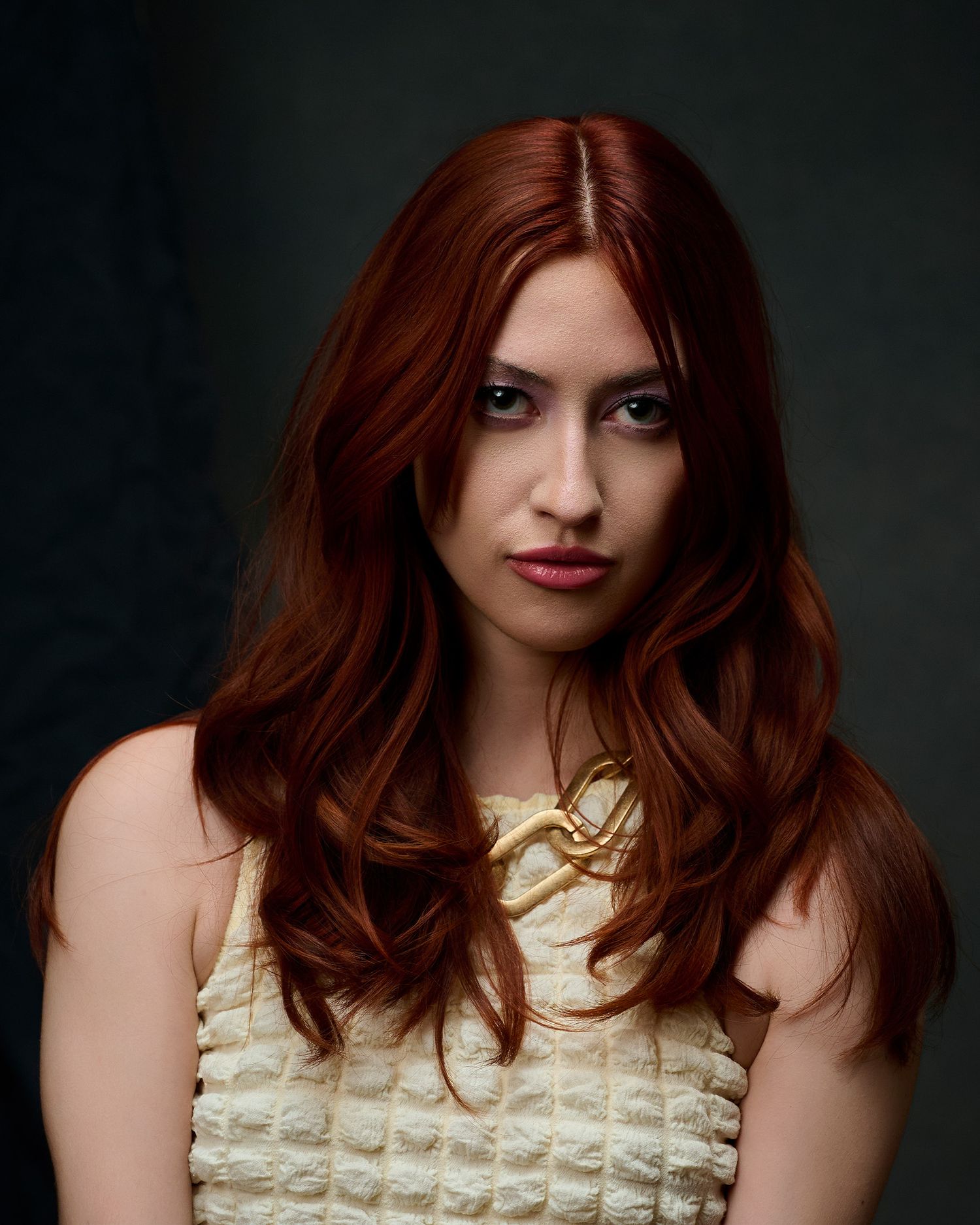 A woman with long, wavy auburn red hair wears a cream textured top in a dramatic studio portrait.