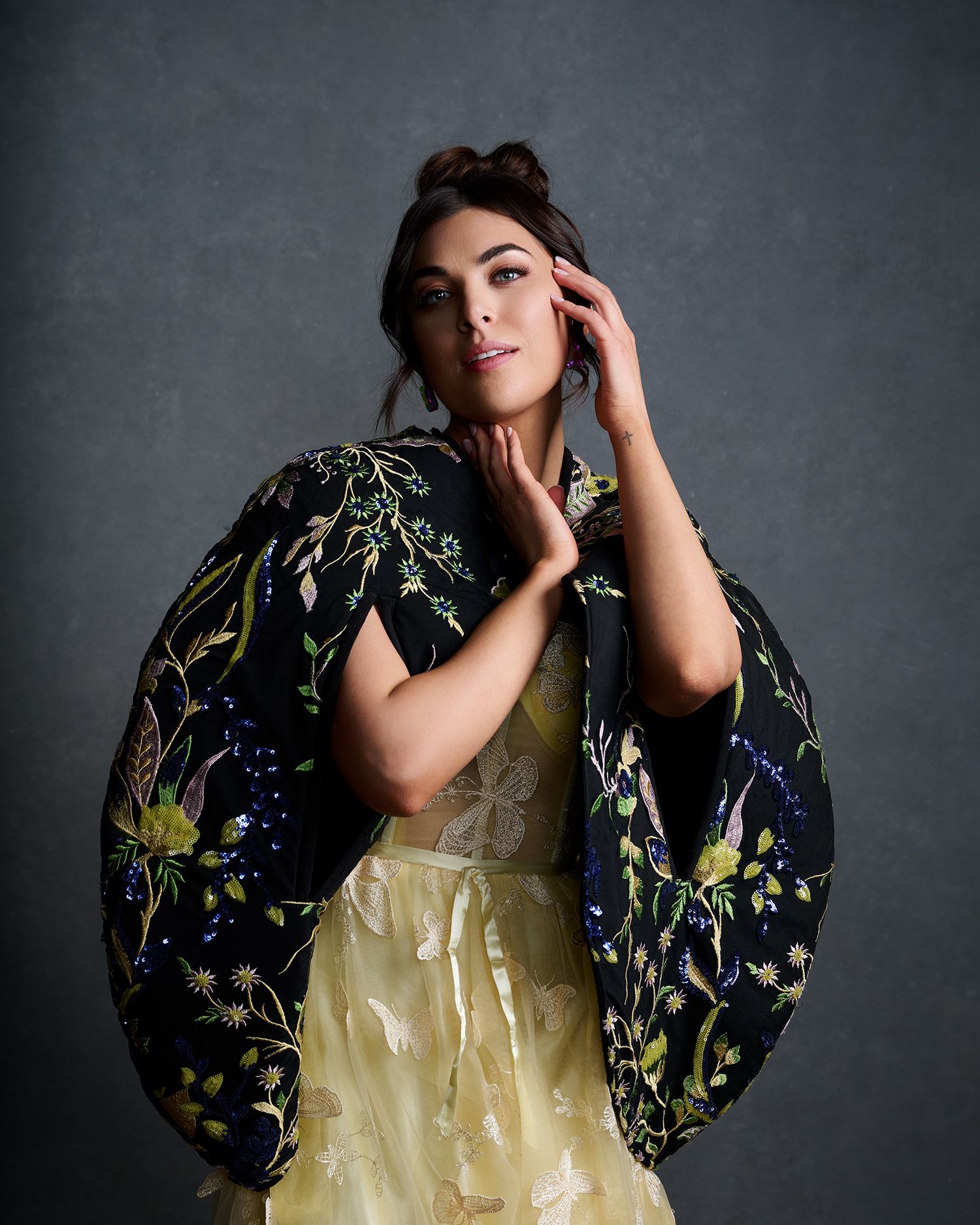 A woman poses gracefully in a floral embroidered black cape over a yellow gown in a studio setting.