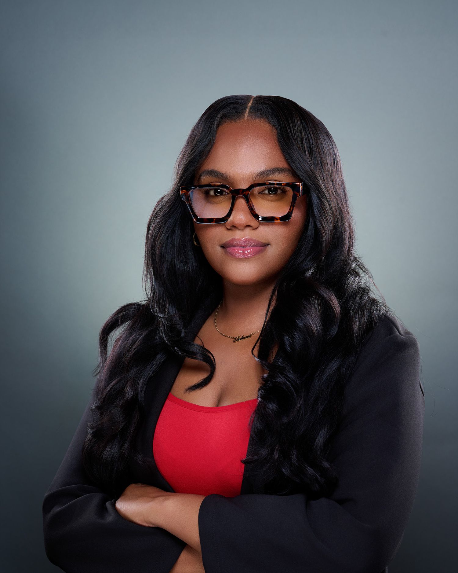Professional headshot of a young woman in glasses and red top with black blazer, arms crossed.