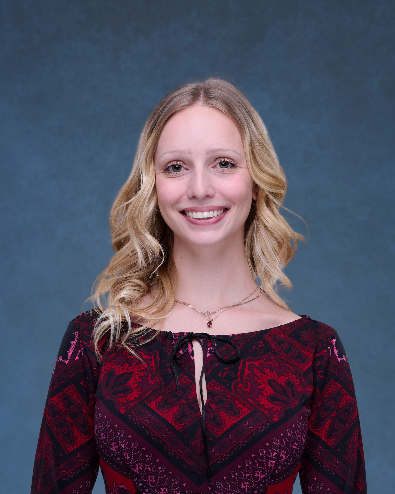 Smiling young blonde woman in red patterned dress poses for professional headshot photo against blue-gray background.