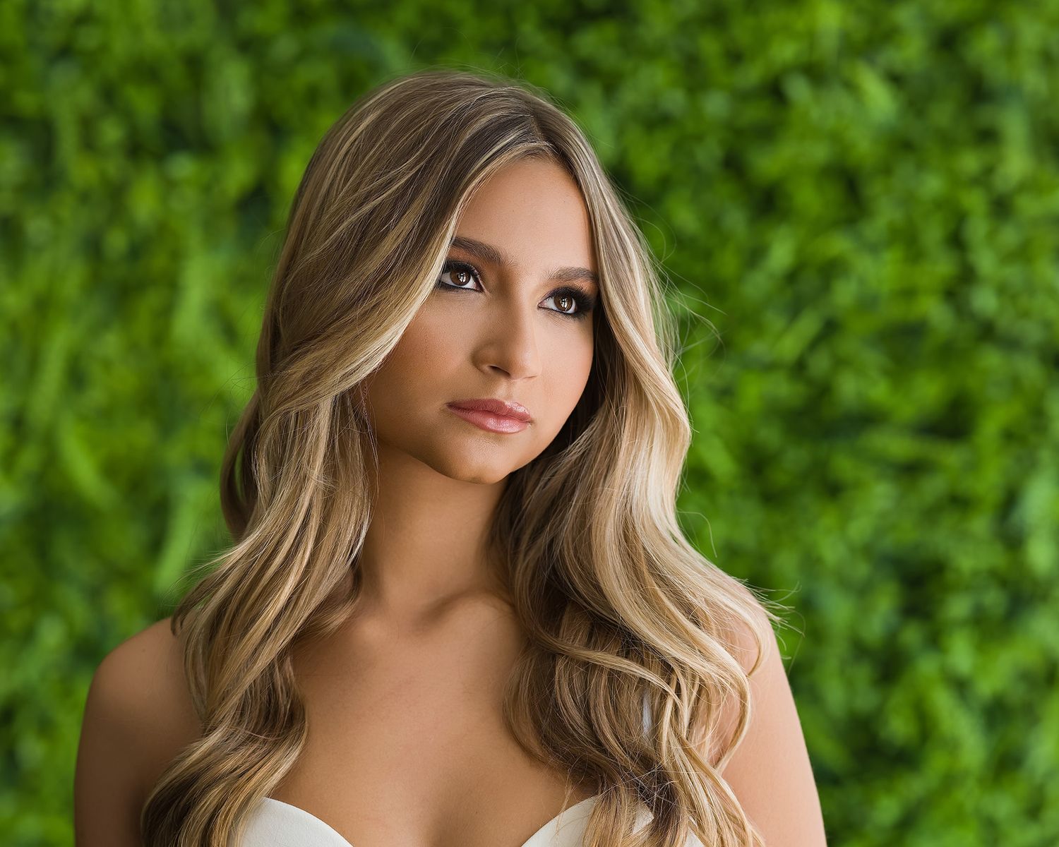 Beautiful young woman with long blonde highlighted waves poses in front of lush green foliage backdrop.