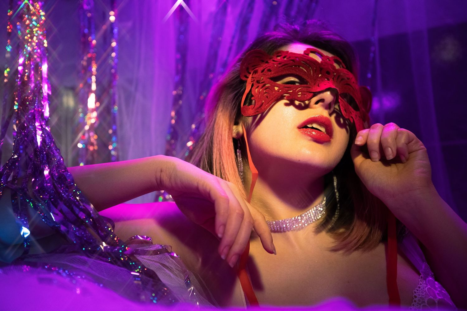 Editorial fashion portrait of a female model wearing a red masquerade mask under purple cinematic lighting.