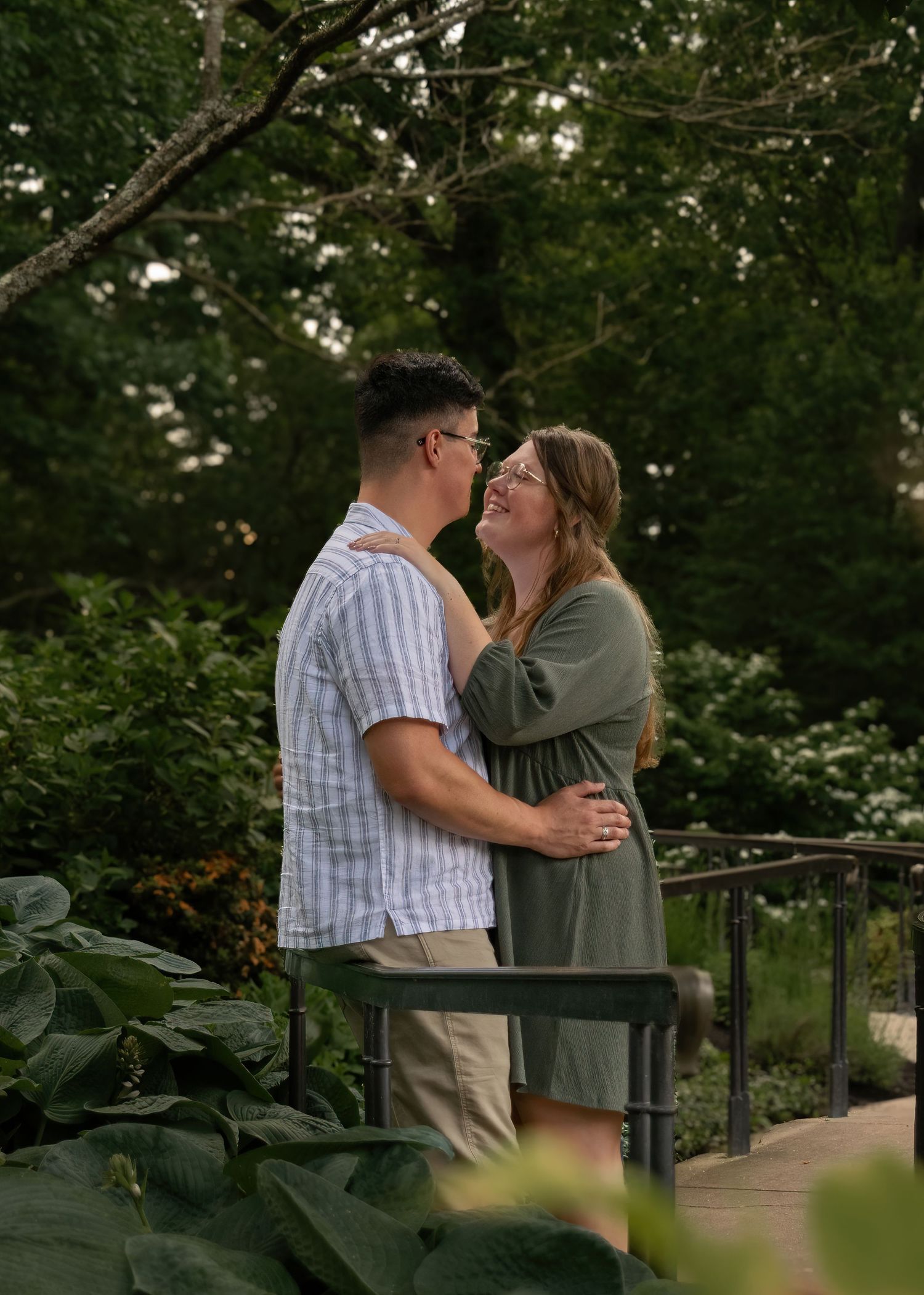 Smiling couple embracing on garden bridge surrounded by lush green foliage.
