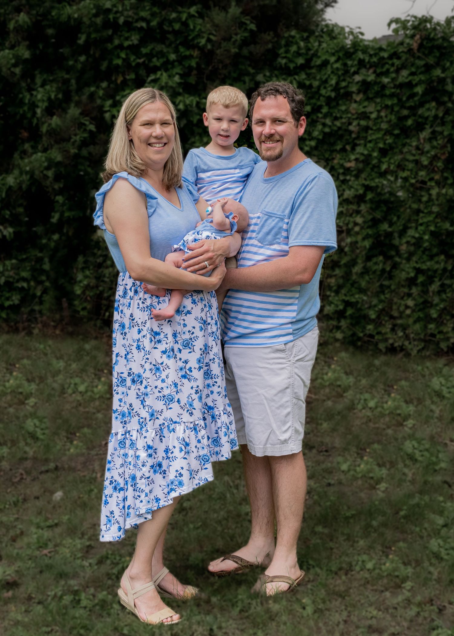 Smiling family of four in matching blue outfits posing outdoors with toddler and newborn baby.