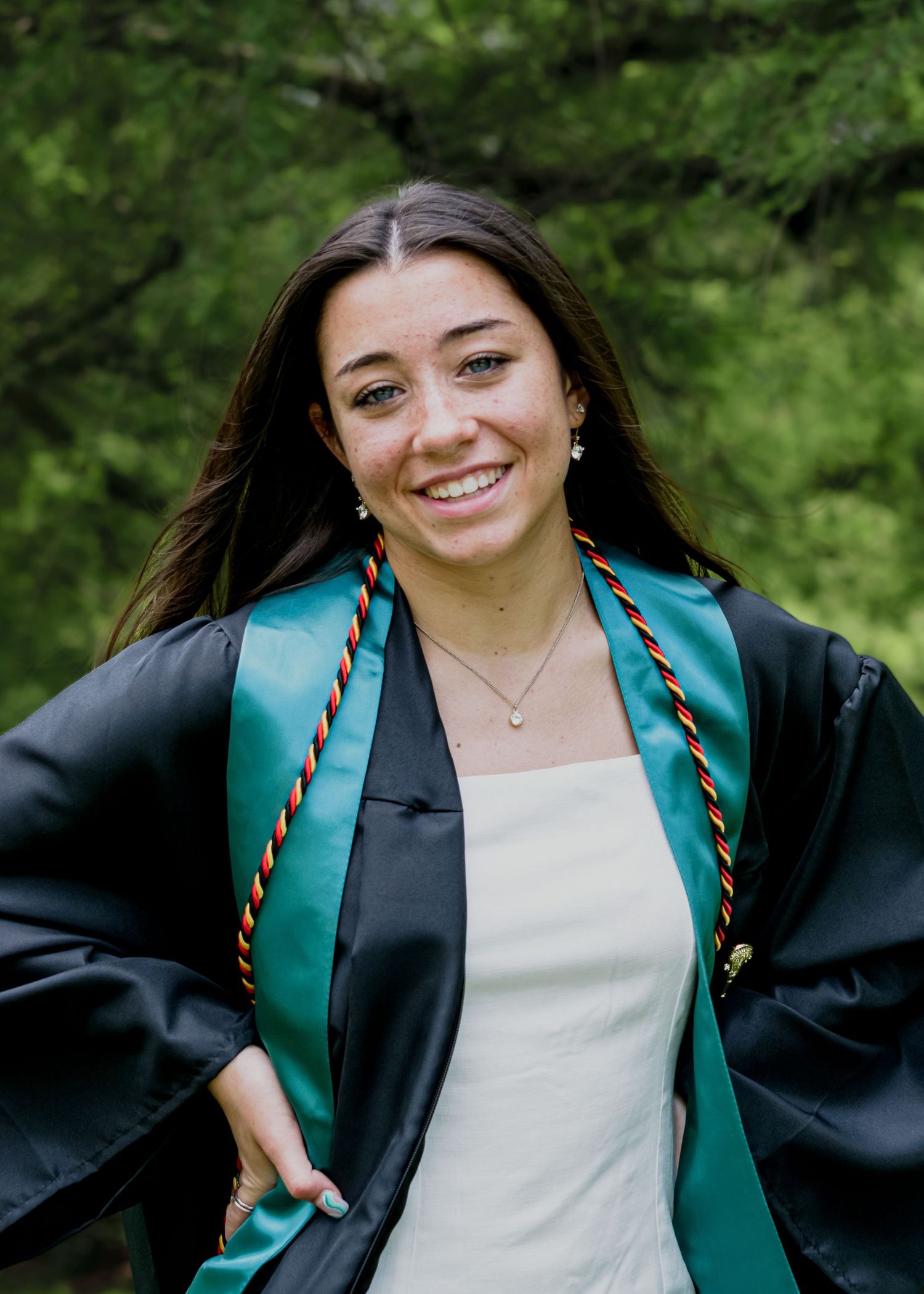 Smiling graduate in black and green graduation gown with honor cords posing outdoors.