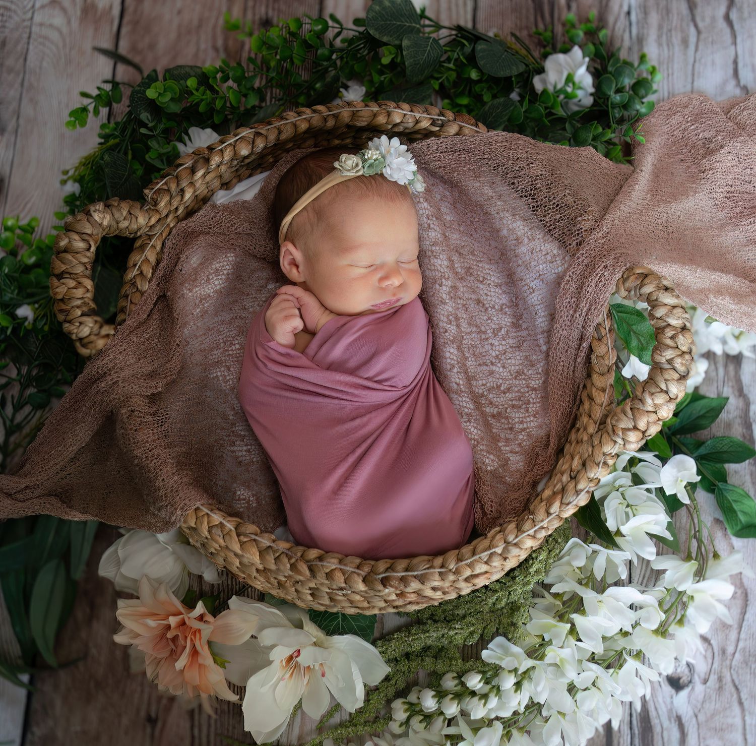Overhead view of sleeping newborn in pink swaddle with floral headband in wicker basket with white flowers.