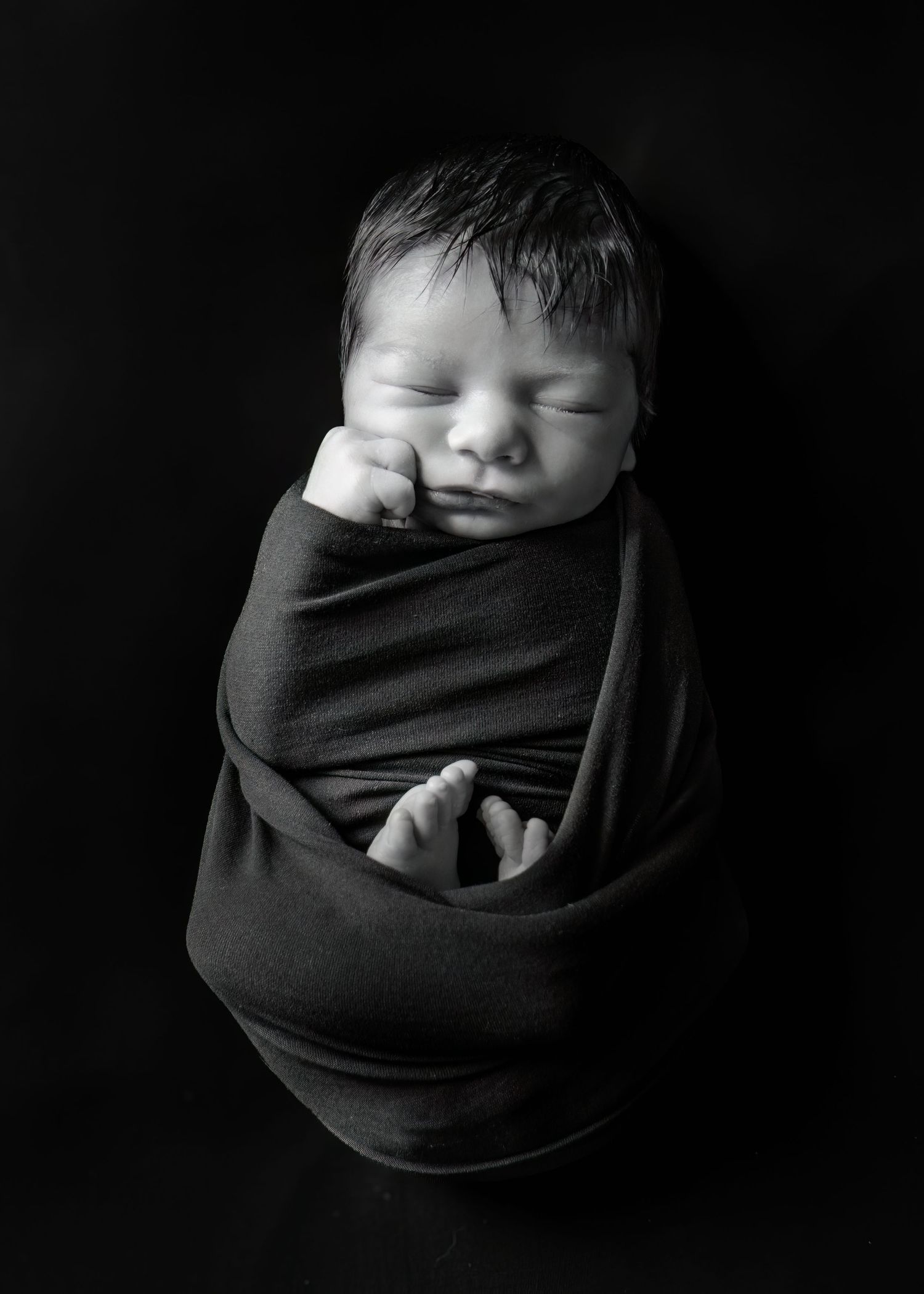 Newborn baby swaddled in dark wrap, black and white portrait photography.