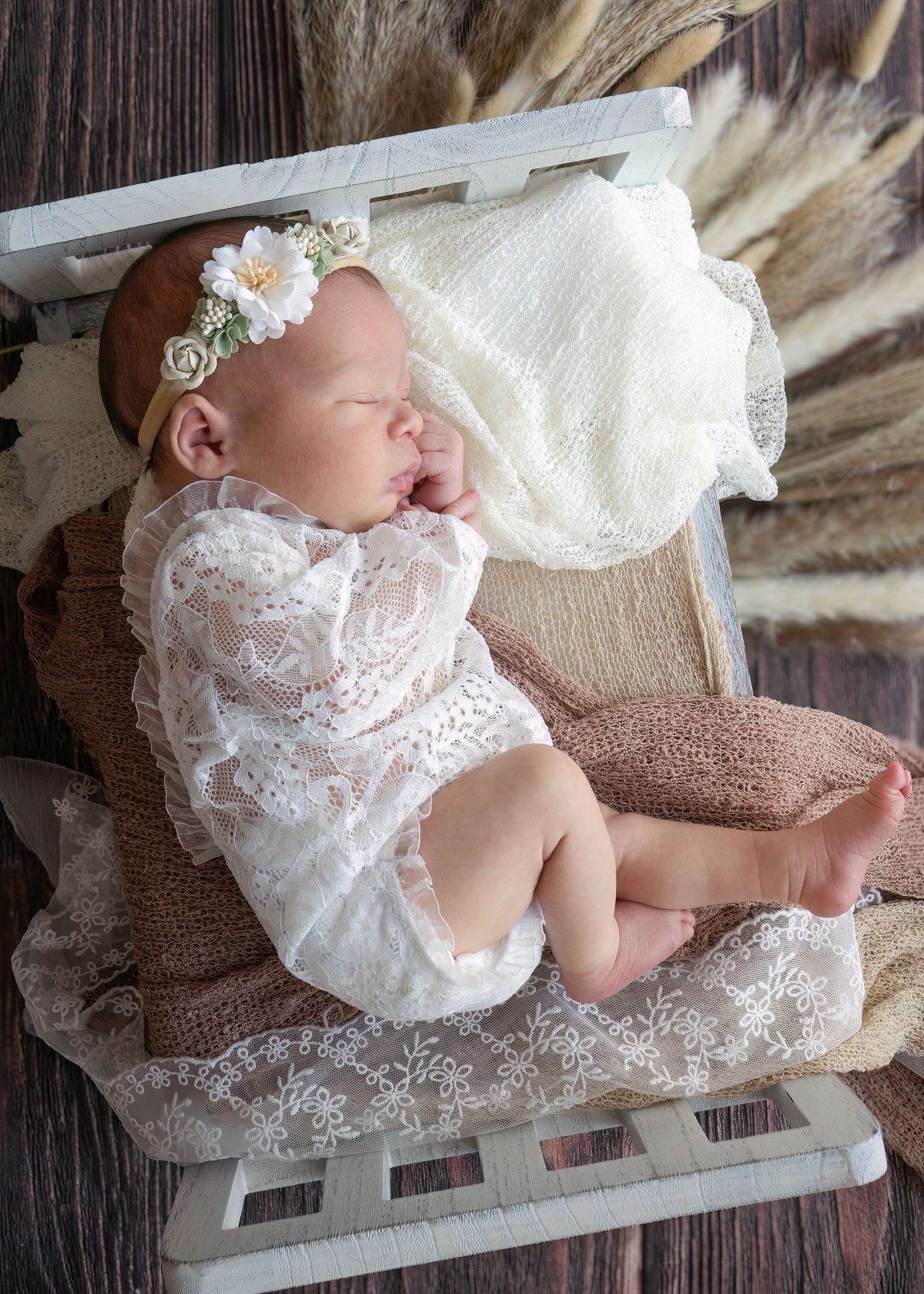 Sleeping newborn baby in white lace outfit with floral headband on rustic wooden prop.
