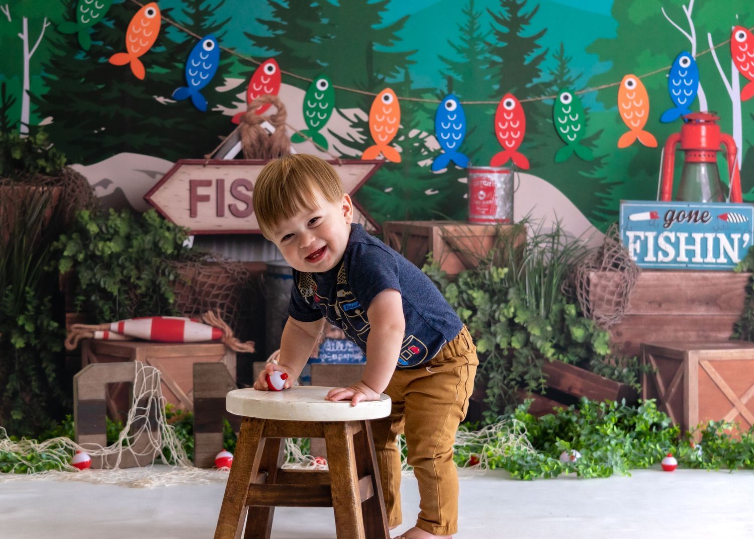 Happy toddler boy smiles and leans on stool at colorful fishing-themed birthday photography session.