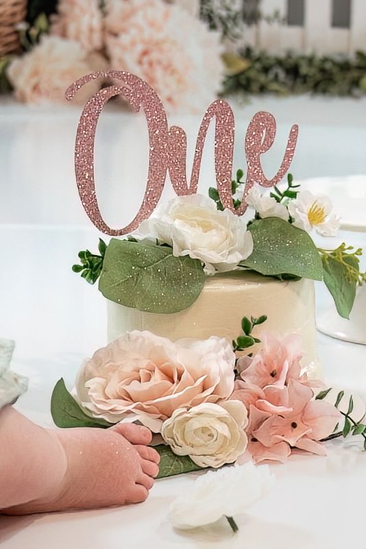 Close-up of white first birthday smash cake with pink roses and glitter One topper with baby feet visible.