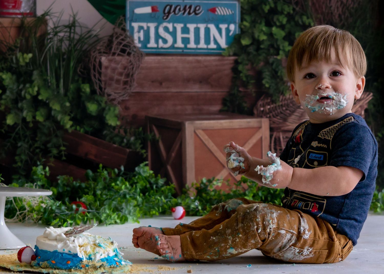 Baby boy with cake smash frosting on face sitting in front of fishing themed backdrop and cake.