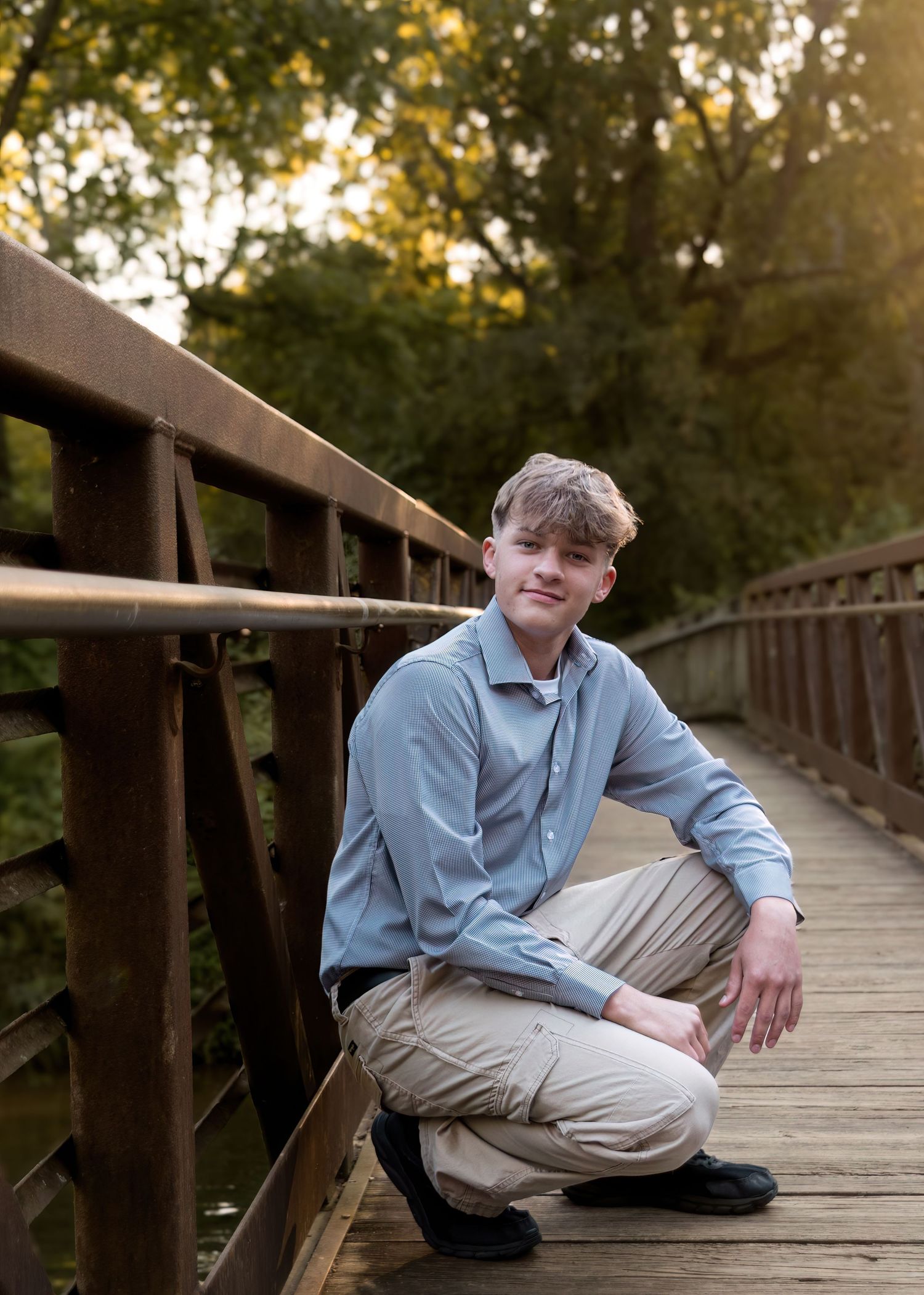 Young man in blue shirt crouching on wooden bridge surrounded by autumn trees.