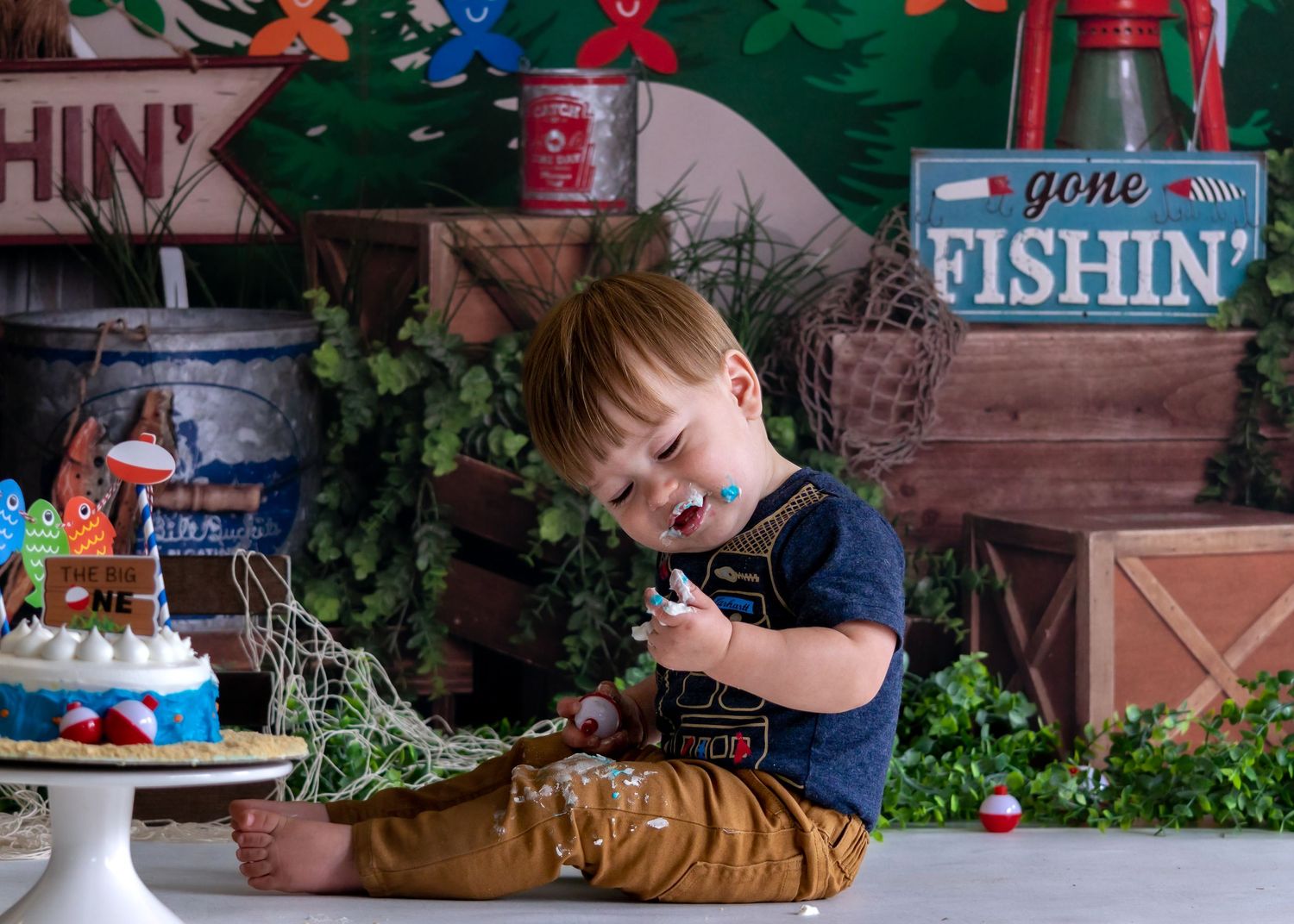 Toddler boy laughing at gone fishing themed cake smash with decorative net and fishing props.
