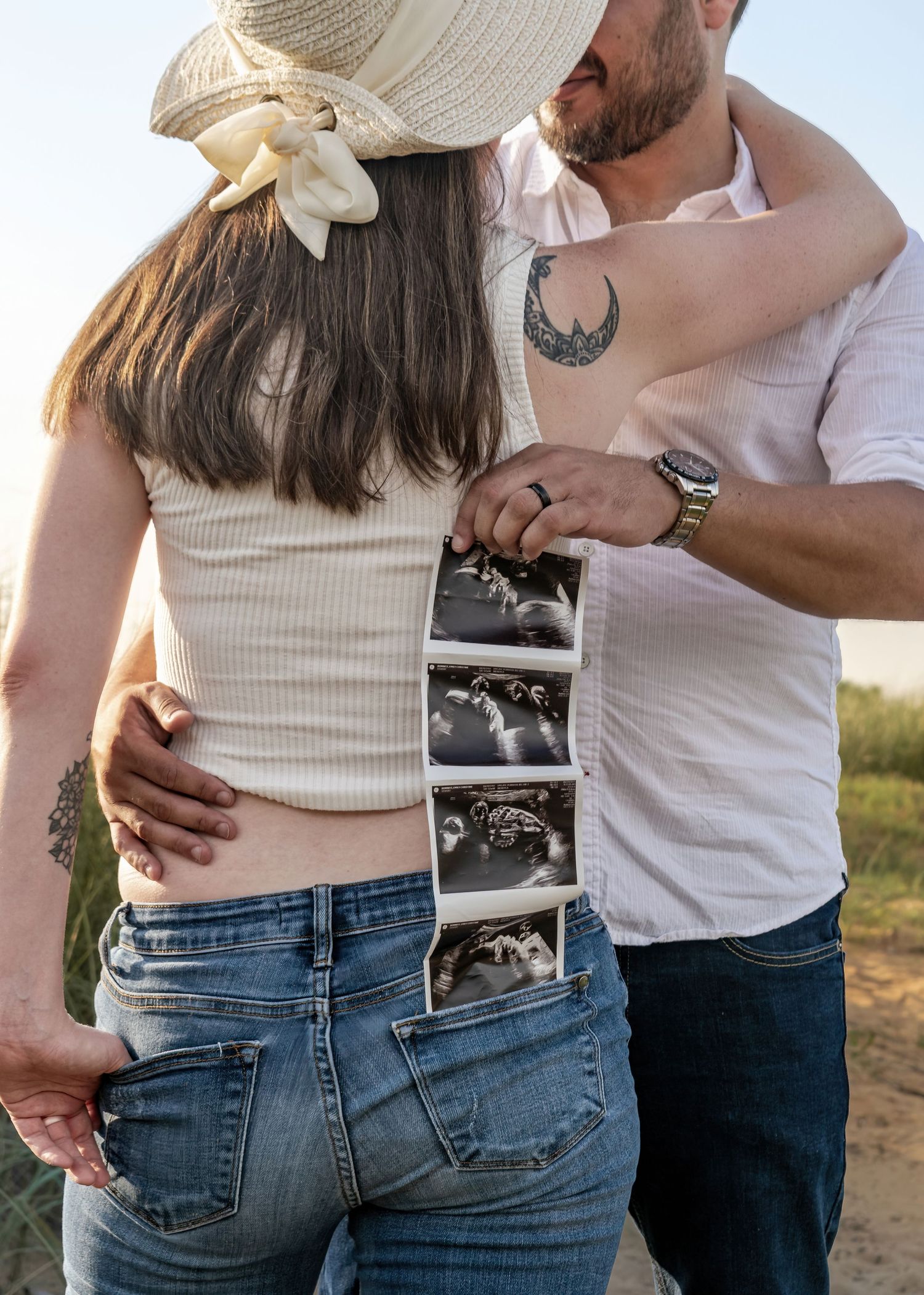 Couple embracing outdoors while man holds ultrasound strip photos behind pregnant woman's back.