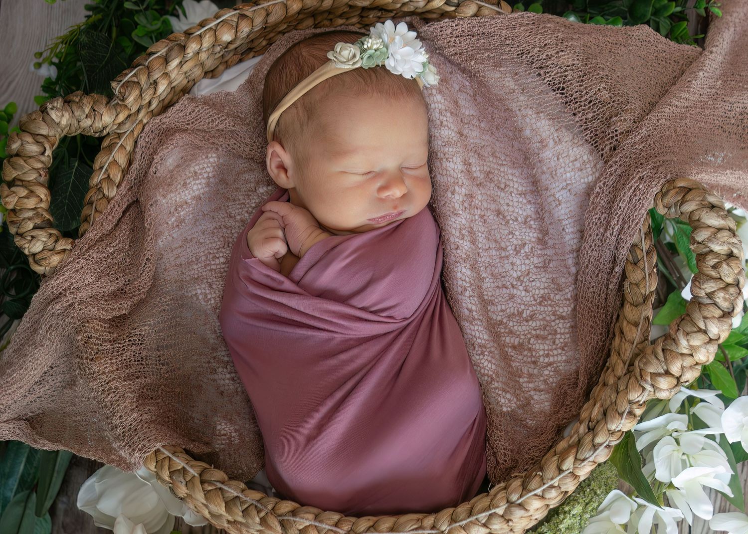Peaceful newborn baby swaddled in mauve wrap with white flower headband resting in wicker basket.
