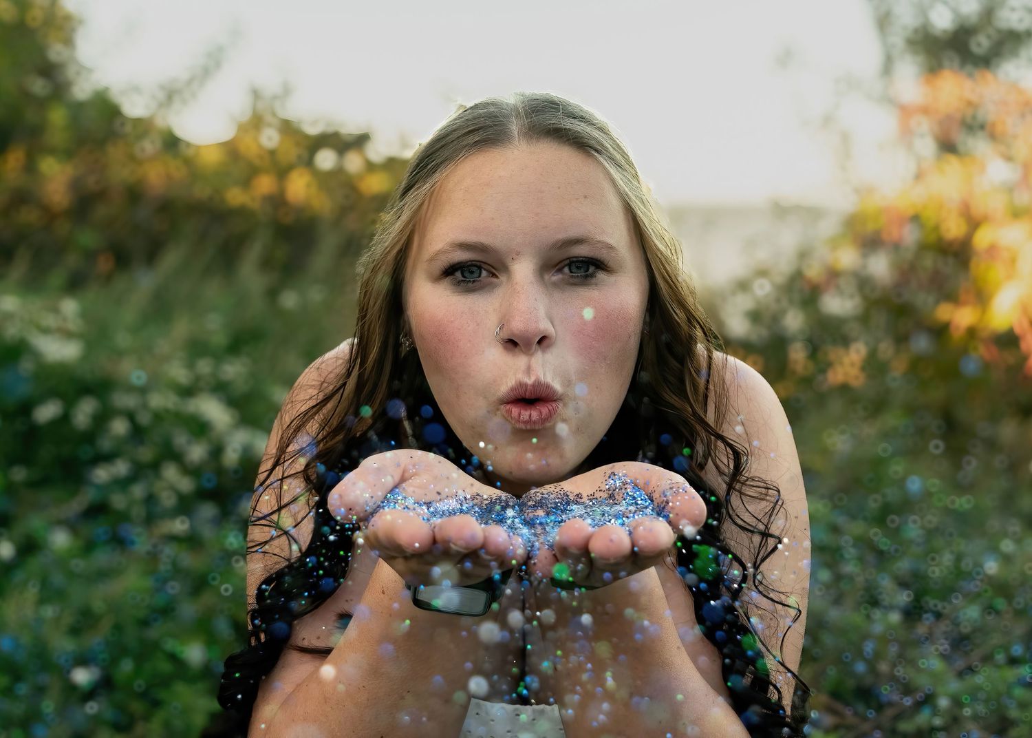 Young woman blowing blue glitter from her hands in a colorful outdoor garden setting.