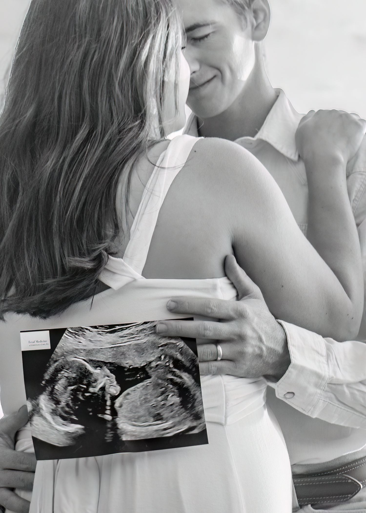 Black and white photo of a couple embracing while holding an ultrasound photo, pregnancy announcement.